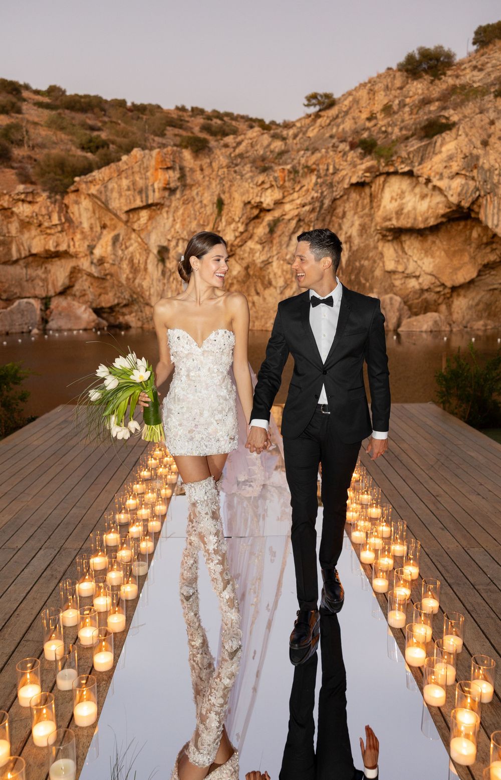 Newlyweds walk down candlelit aisle on wooden dock with stunning rocky cliffs behind them.