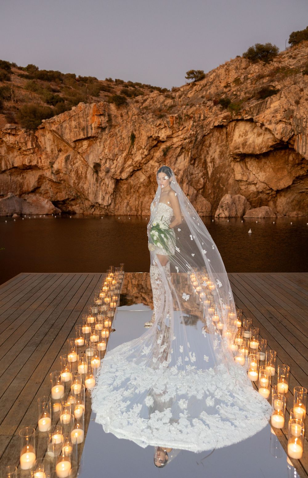 A dramatic wedding dress and veil illuminated by candlelight against rocky cliffs at dusk.