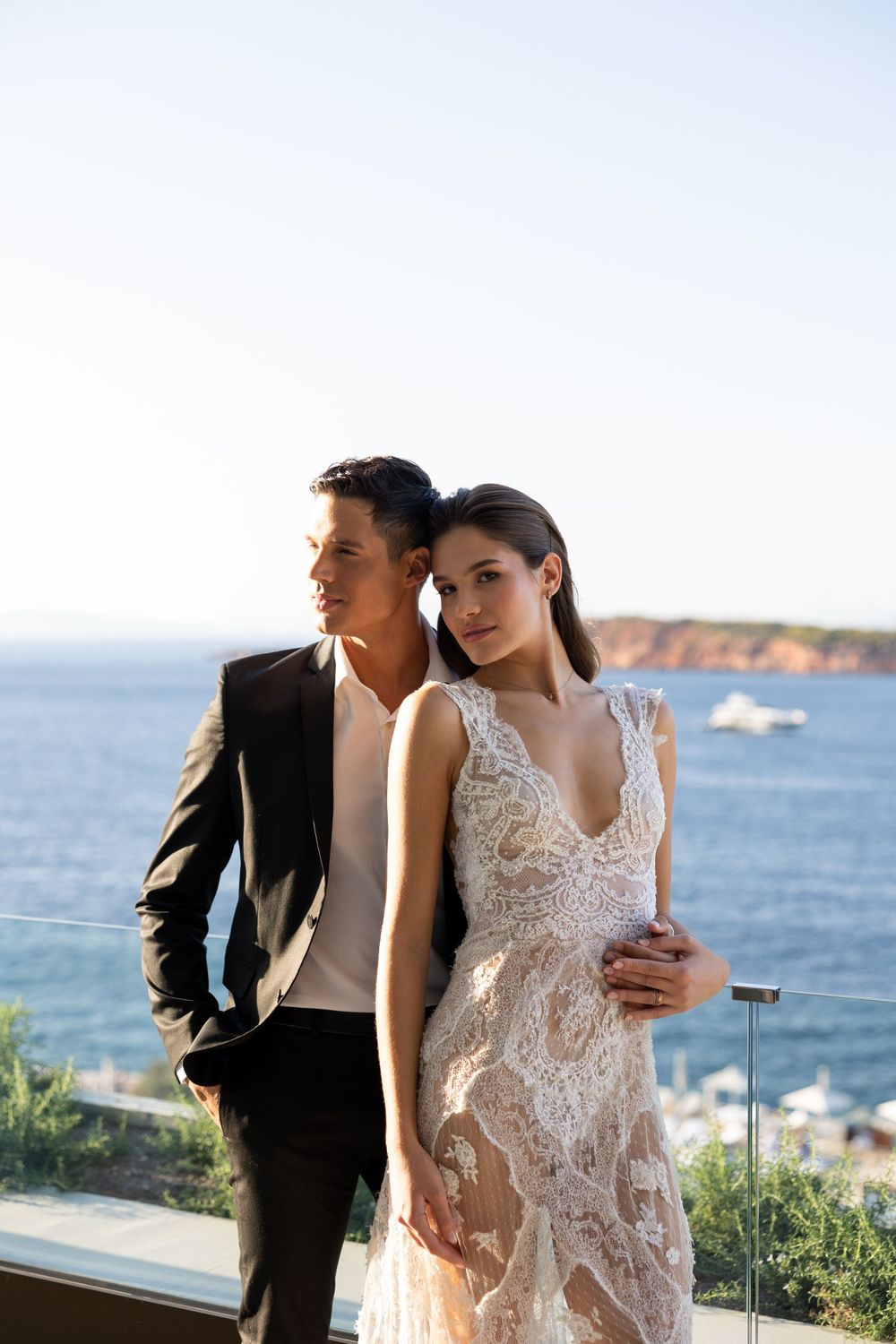 A couple poses against a coastal backdrop with cliffs and ocean, the bride wearing an elegant lace dress.