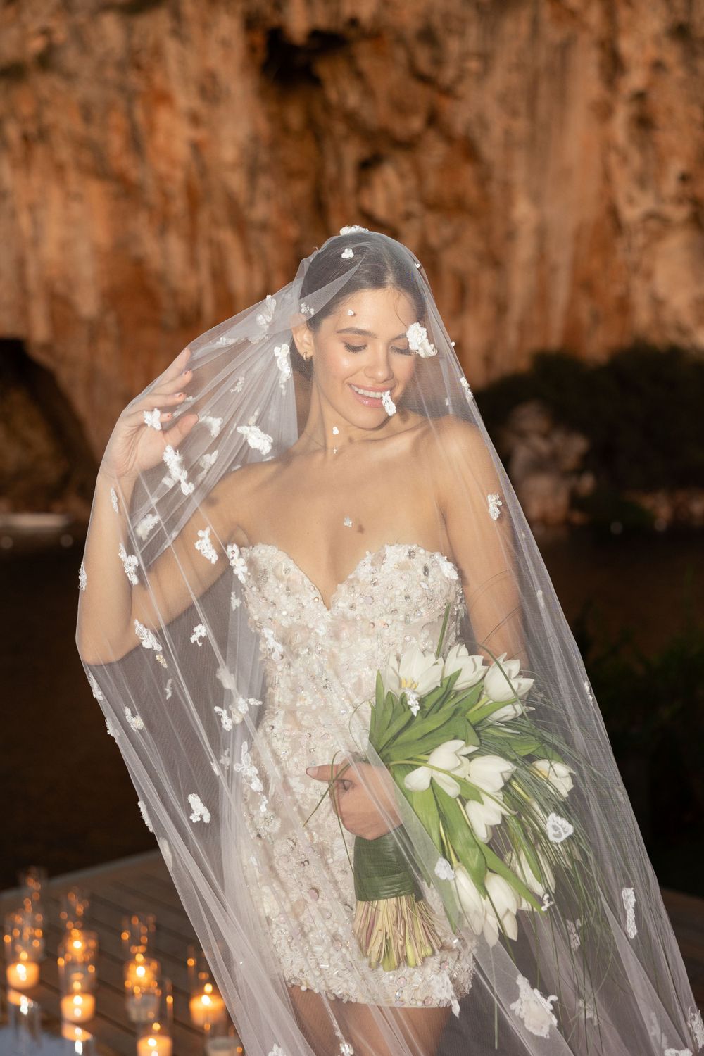 A bride in a strapless lace dress holds a white calla lily bouquet while wearing an embellished cathedral veil.