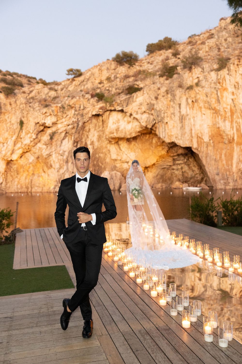 A luxury wedding ceremony setup on a wooden deck against dramatic cliffs with glowing candles lining the pathway at sunset.
