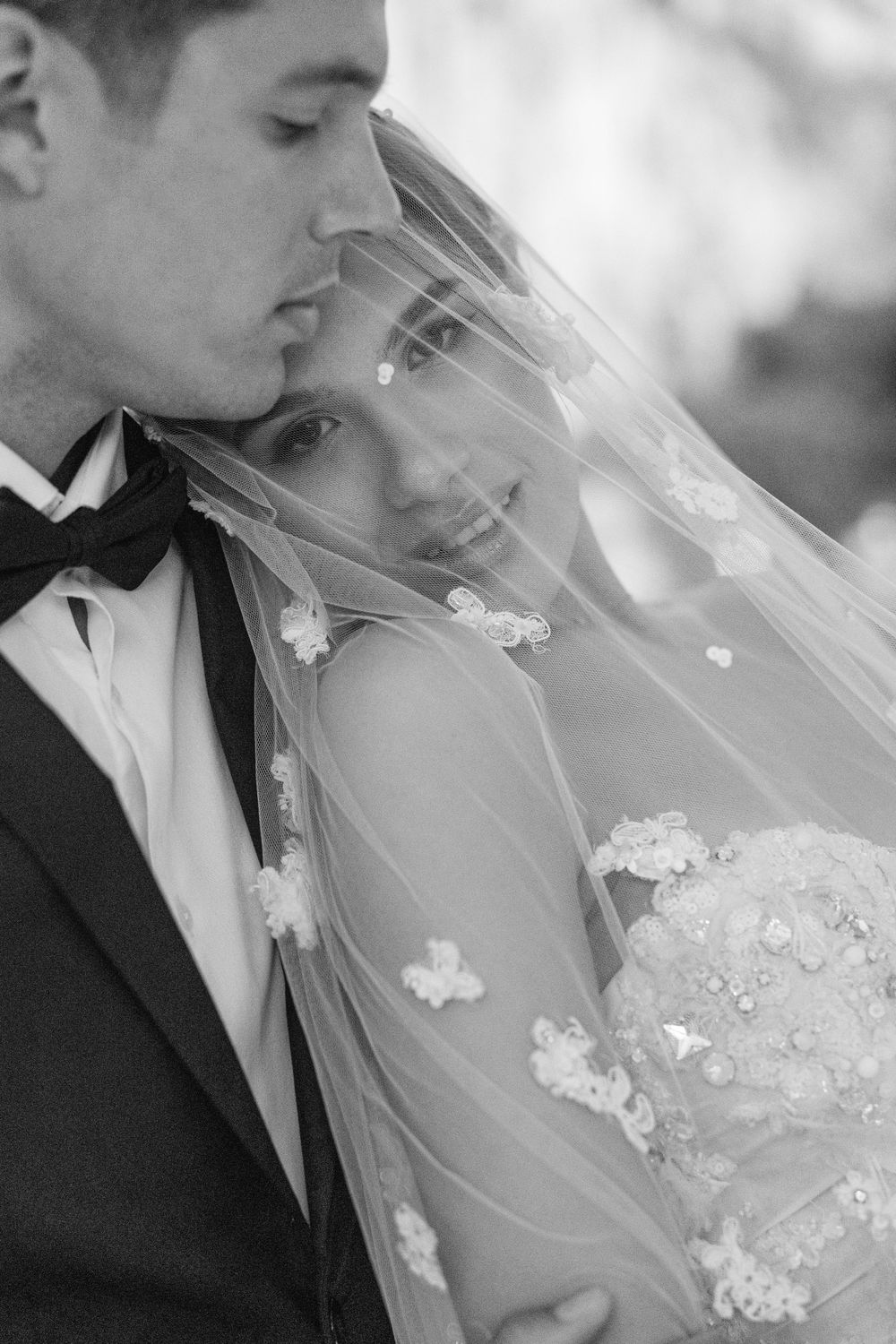 A romantic black and white wedding portrait showing a tender moment through a delicate veil with floral appliques.