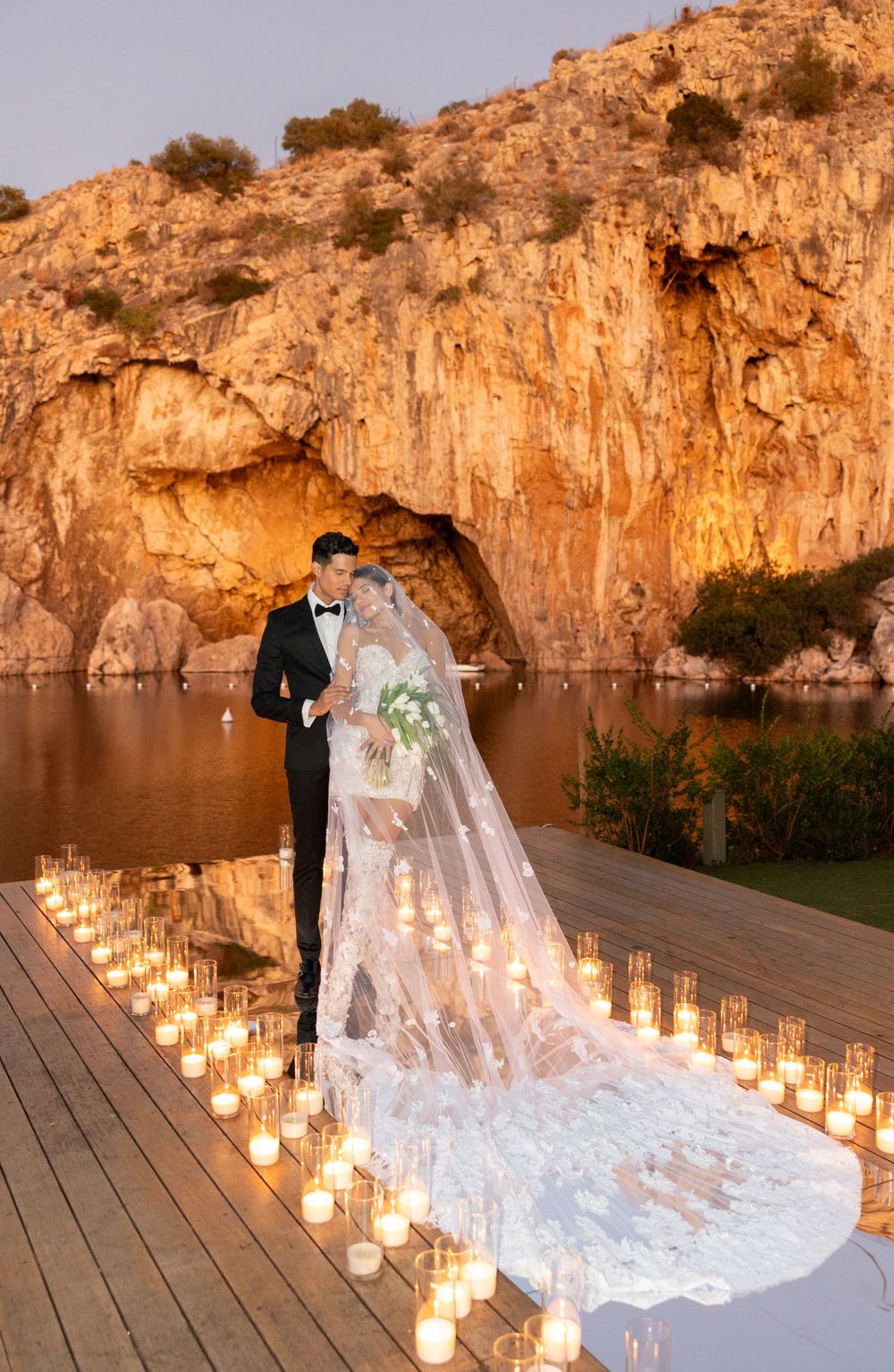 A romantic wedding scene on a wooden dock at sunset with candles lining the path and dramatic cliffside backdrop.