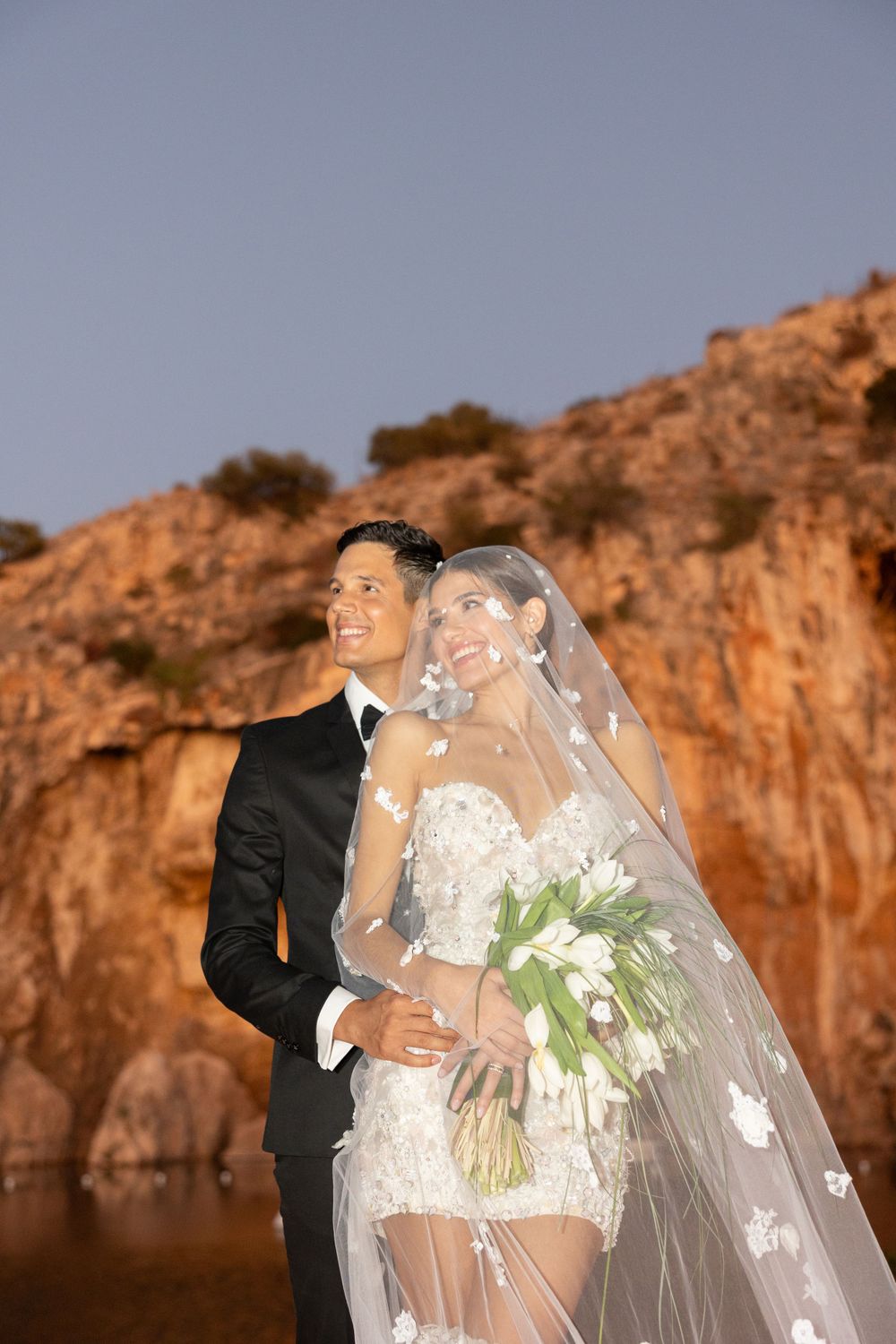 A glamorous wedding couple poses against rocky cliffs at sunset with the bride holding a white floral bouquet.