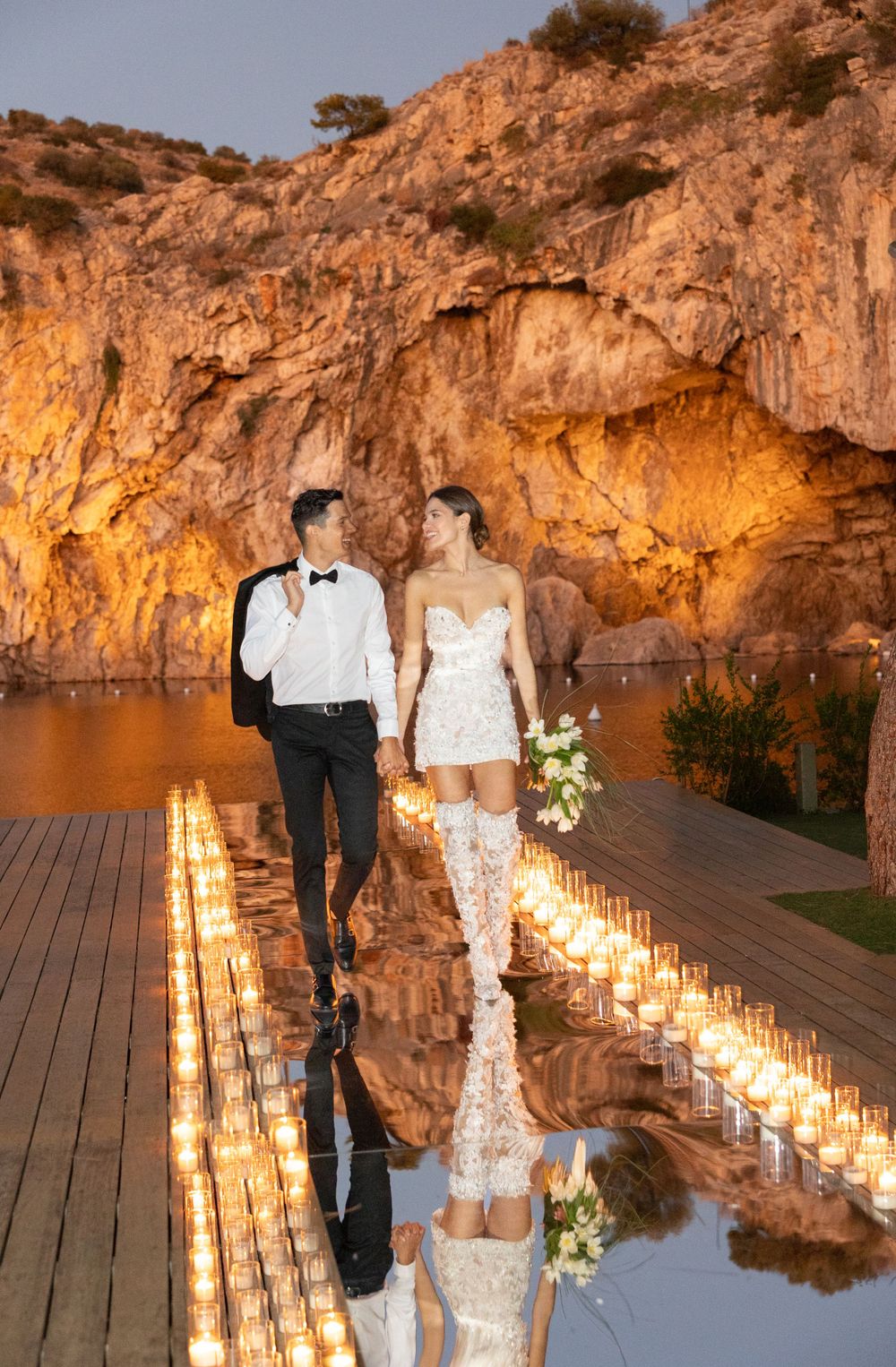 A couple stands on a reflective wooden path lined with candlelight against a dramatic illuminated cliff backdrop.