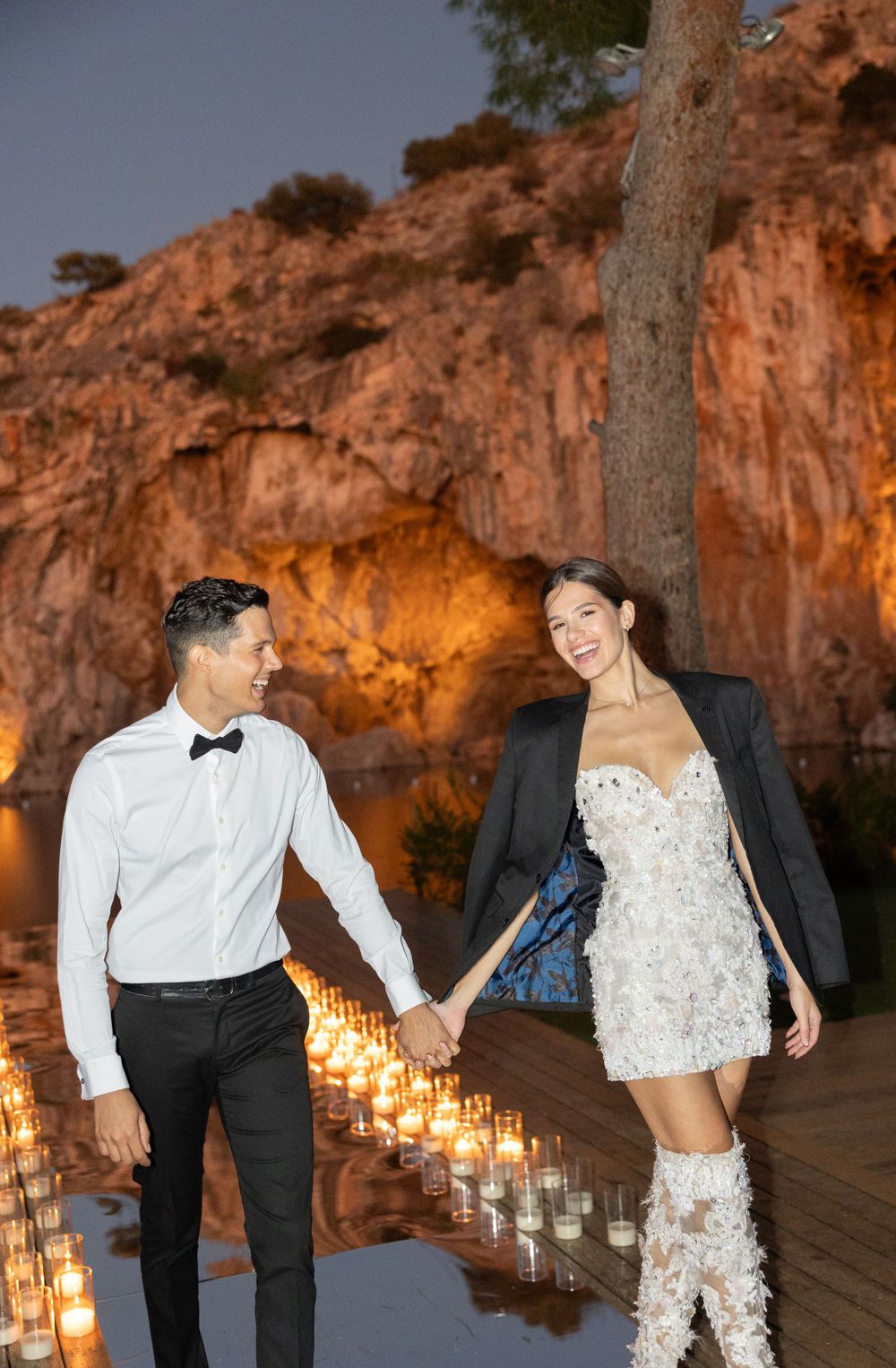 A couple holding hands on a candlelit deck against dramatic red cliffs during a wedding celebration.
