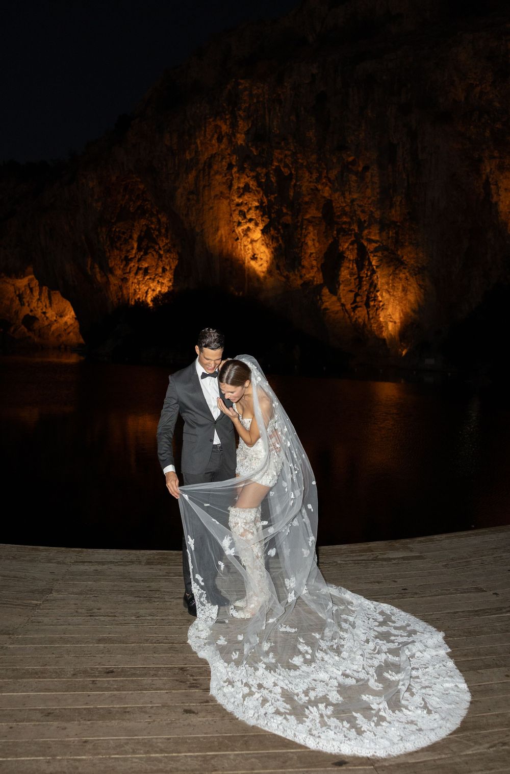 A dramatic nighttime wedding photo with a couple on a dock illuminated against warmly lit cliffs in the background.