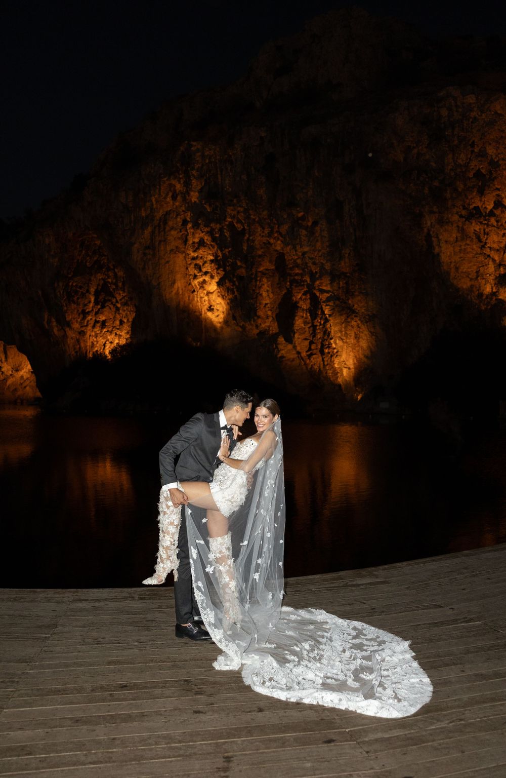 A couple in wedding attire embraces on a wooden dock at night with illuminated cliffs in the background.