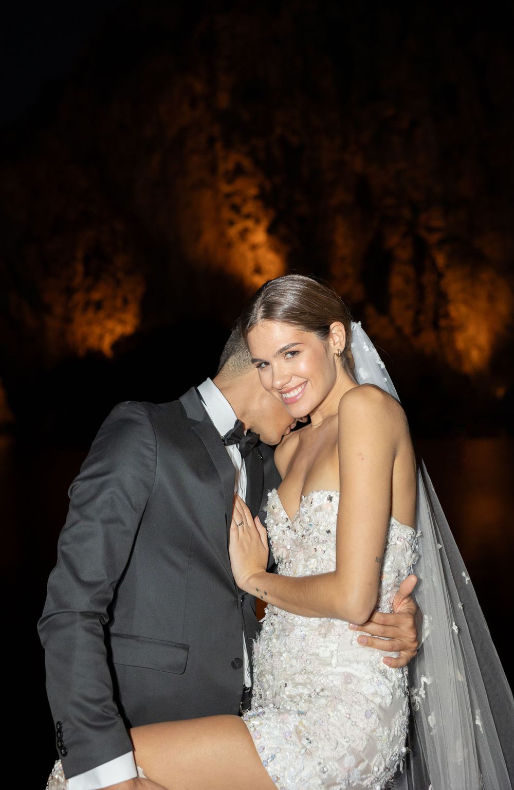 A romantic wedding photo showing a happy couple embracing at night with warm orange lighting in the background.