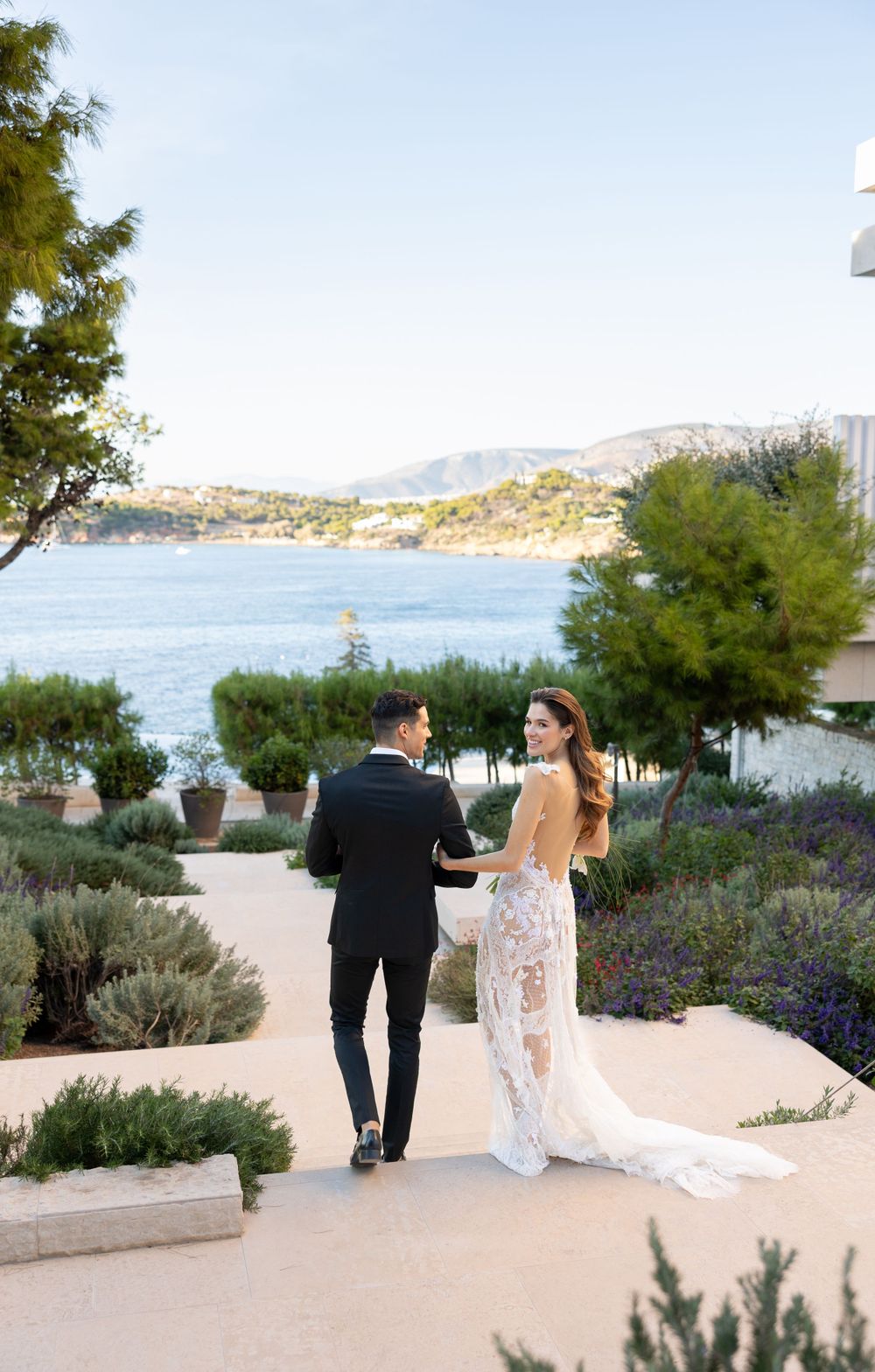 A wedding couple walking on a seaside path with Mediterranean style landscaping and mountains in the background.