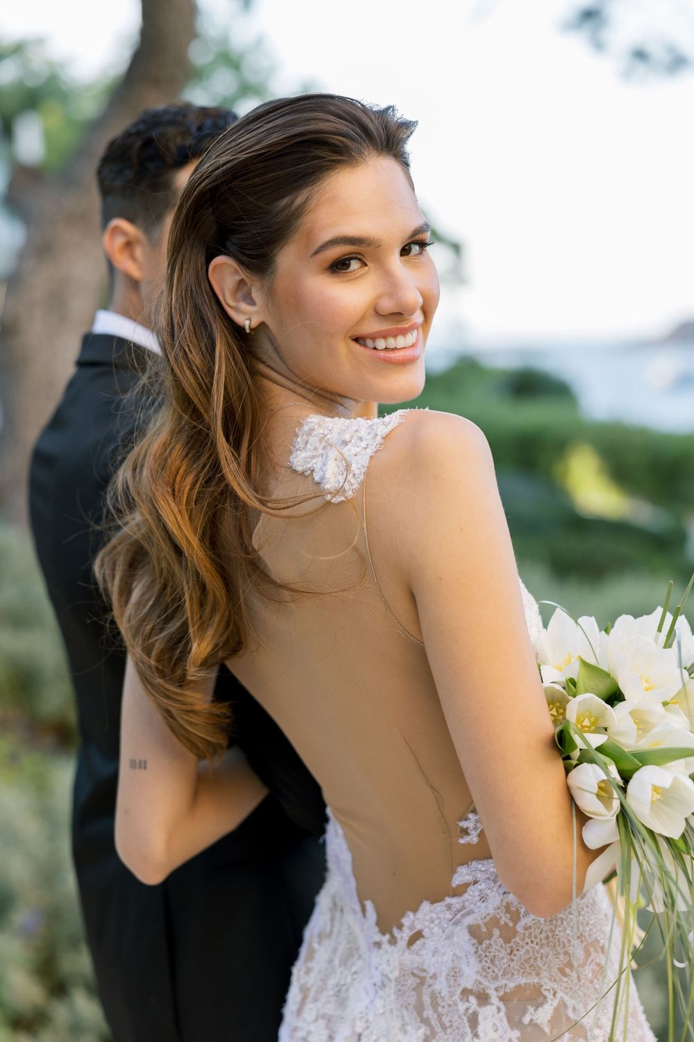 Bride in elegant lace backless wedding dress smiles over her shoulder while holding white bouquet outdoors.