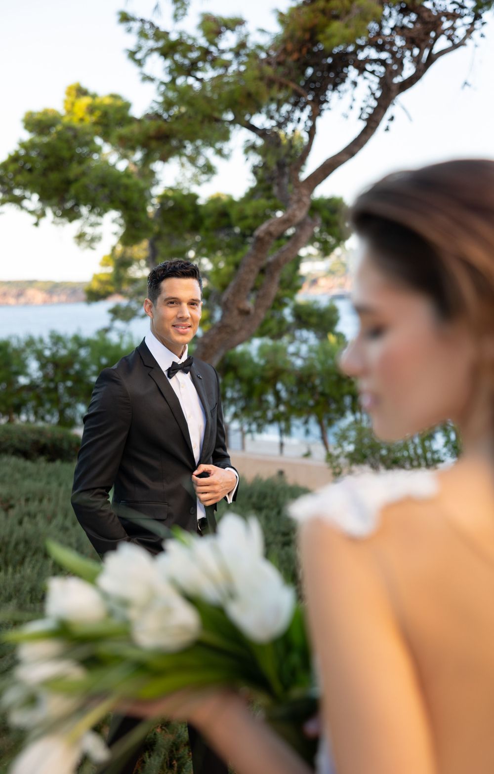 A romantic wedding photo with a couple in formal attire posing by the sea with white flowers in the foreground.
