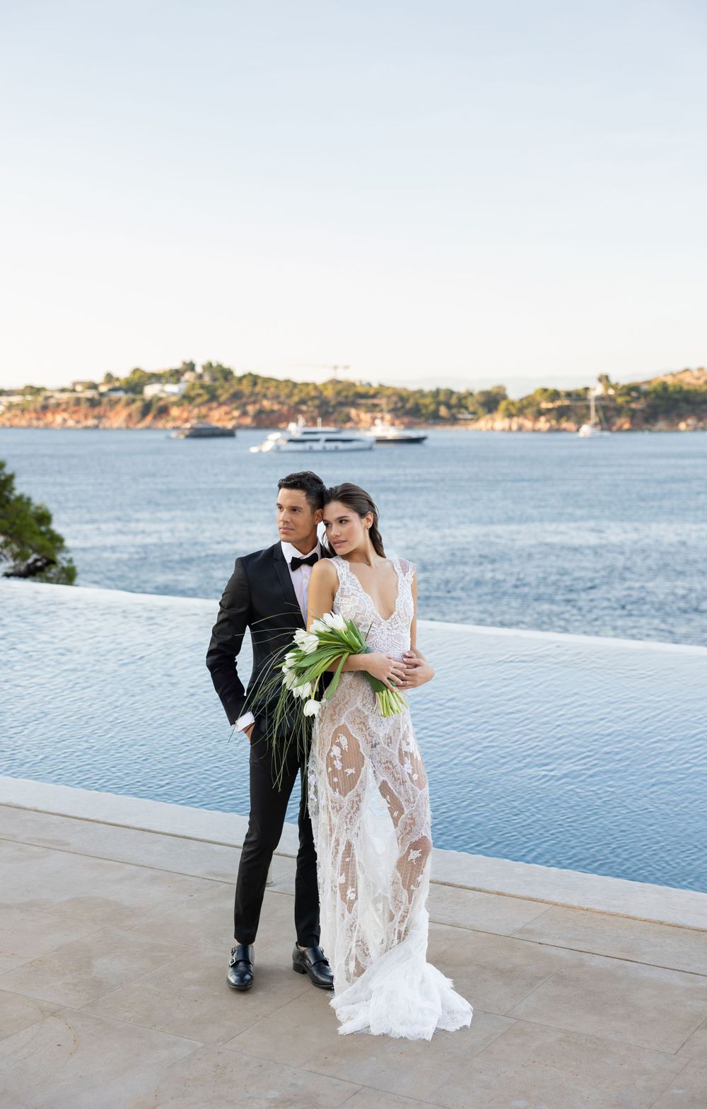 Couple in wedding attire standing by infinity pool overlooking ocean bay with boats in background at sunset.