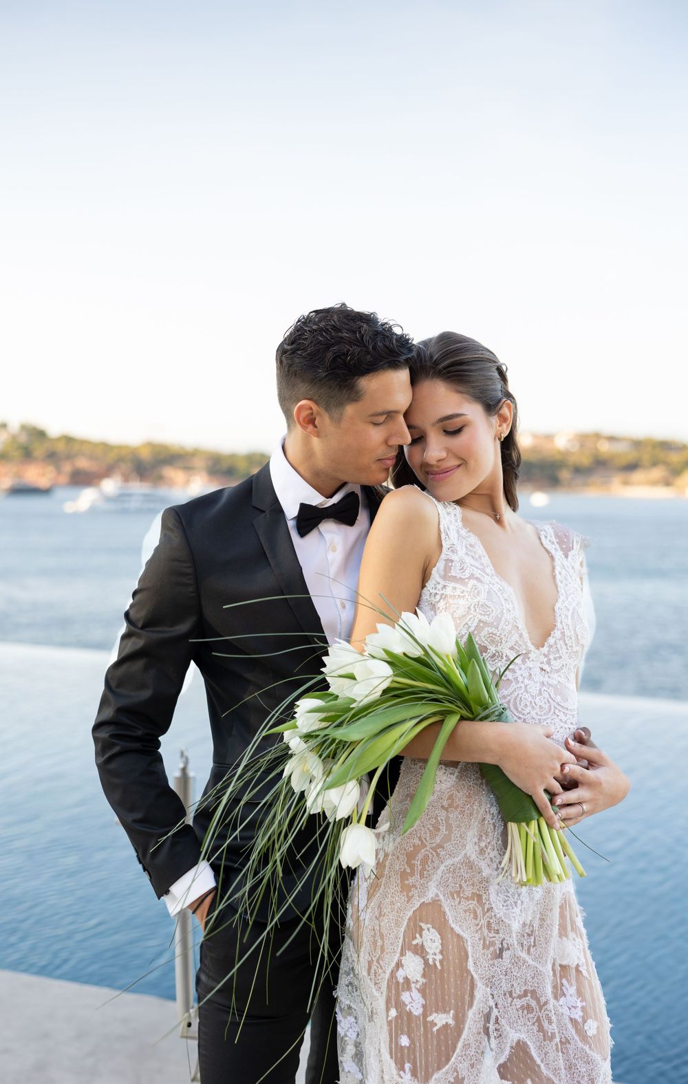 A couple embraces on a waterfront terrace with a bouquet of white lilies.