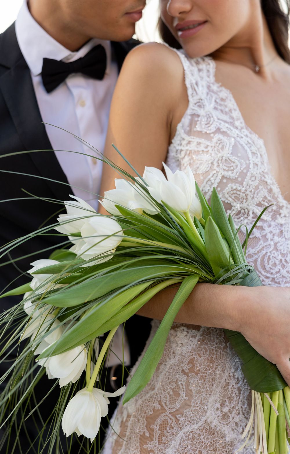 Close-up of newlyweds with white tulip bouquet showcasing intricate lace wedding dress details.