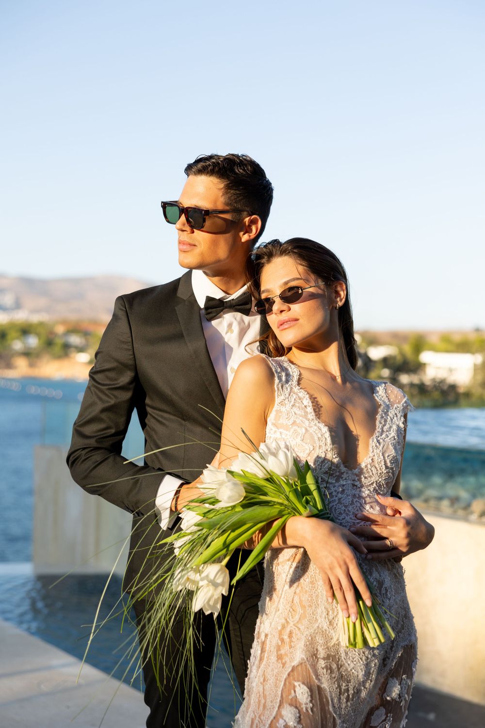 Stylish couple in sunglasses pose on seaside terrace with bride holding white floral bouquet.