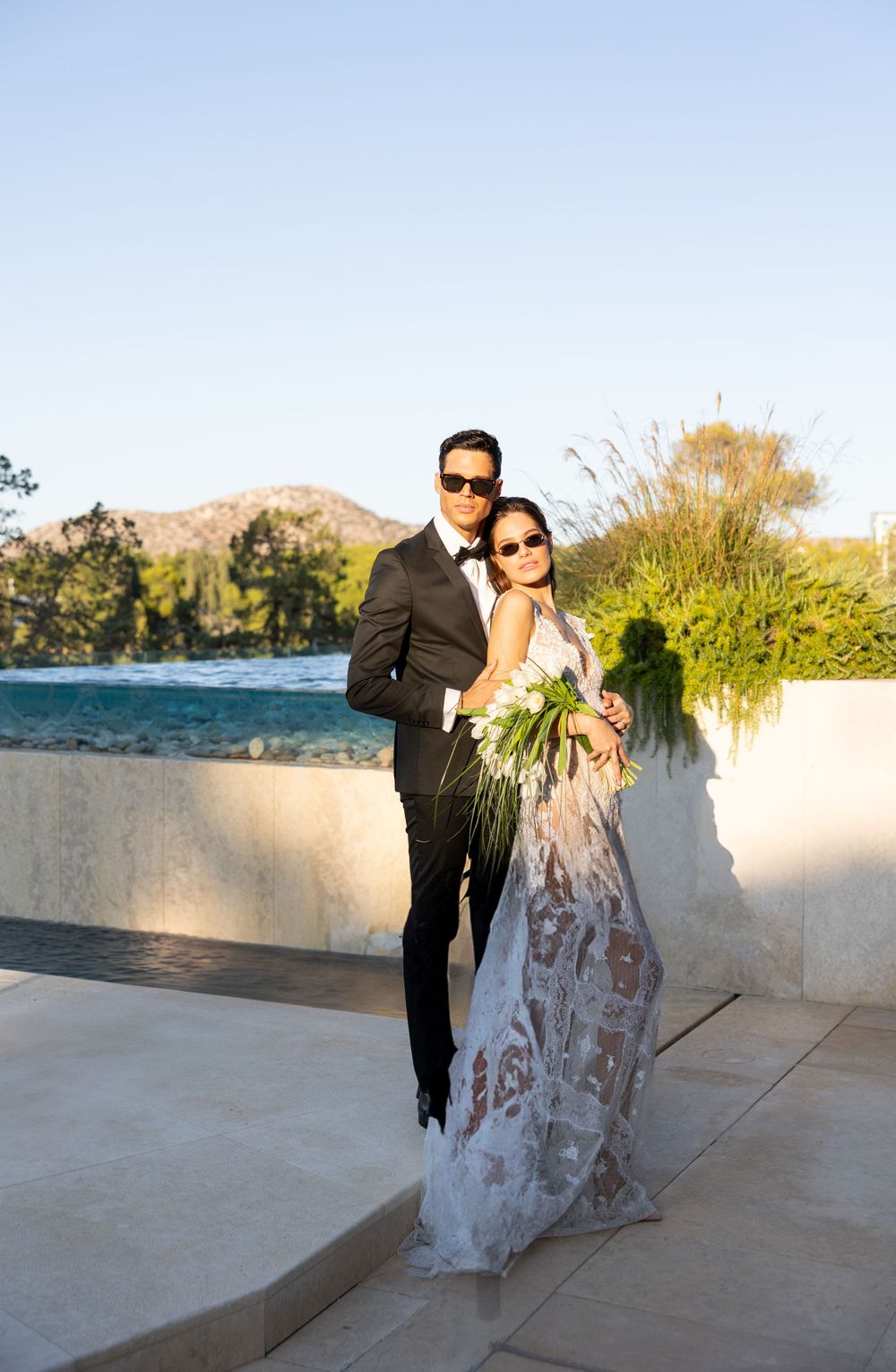 Wedding couple standing on oceanfront terrace with mountains in background during sunset.