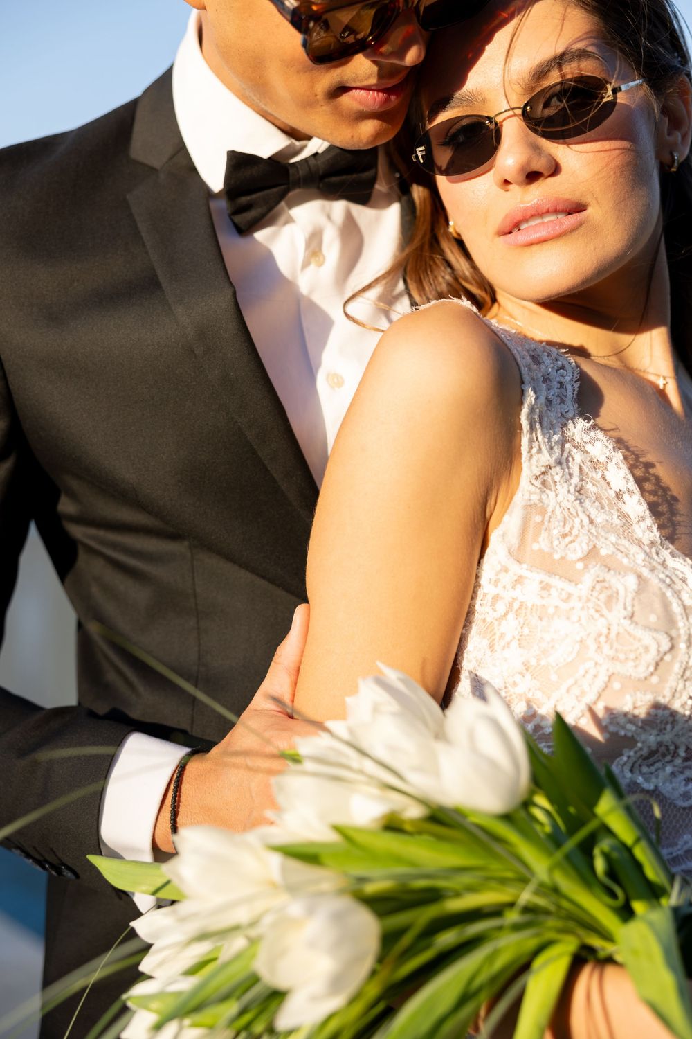 Stylish bride and groom wearing sunglasses pose with white tulips in bright sunlight.