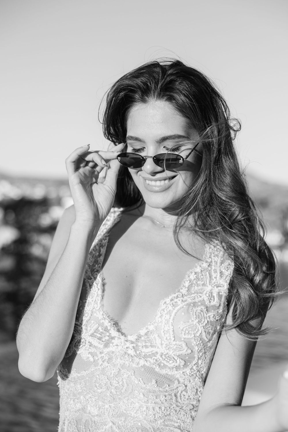 A black and white portrait of a bride in a lace dress adjusting her sunglasses and smiling outdoors.