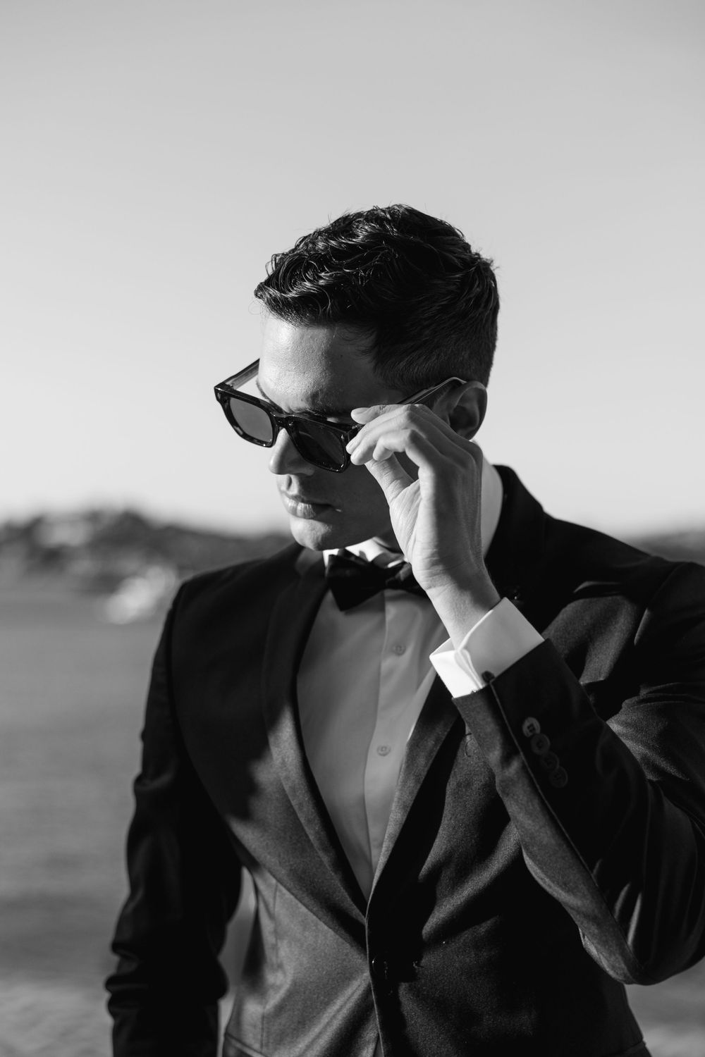 A stylish black and white portrait of a person in a tuxedo and sunglasses adjusting their frames.