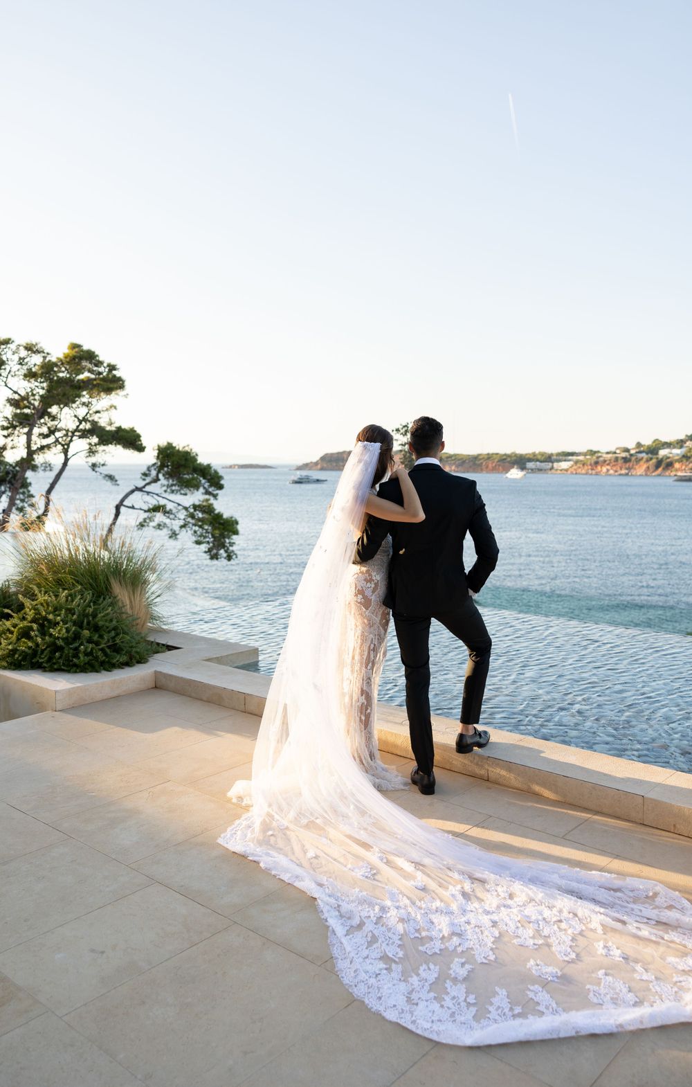 A wedding couple in formal attire admiring the ocean view from a stone terrace overlooking blue waters.