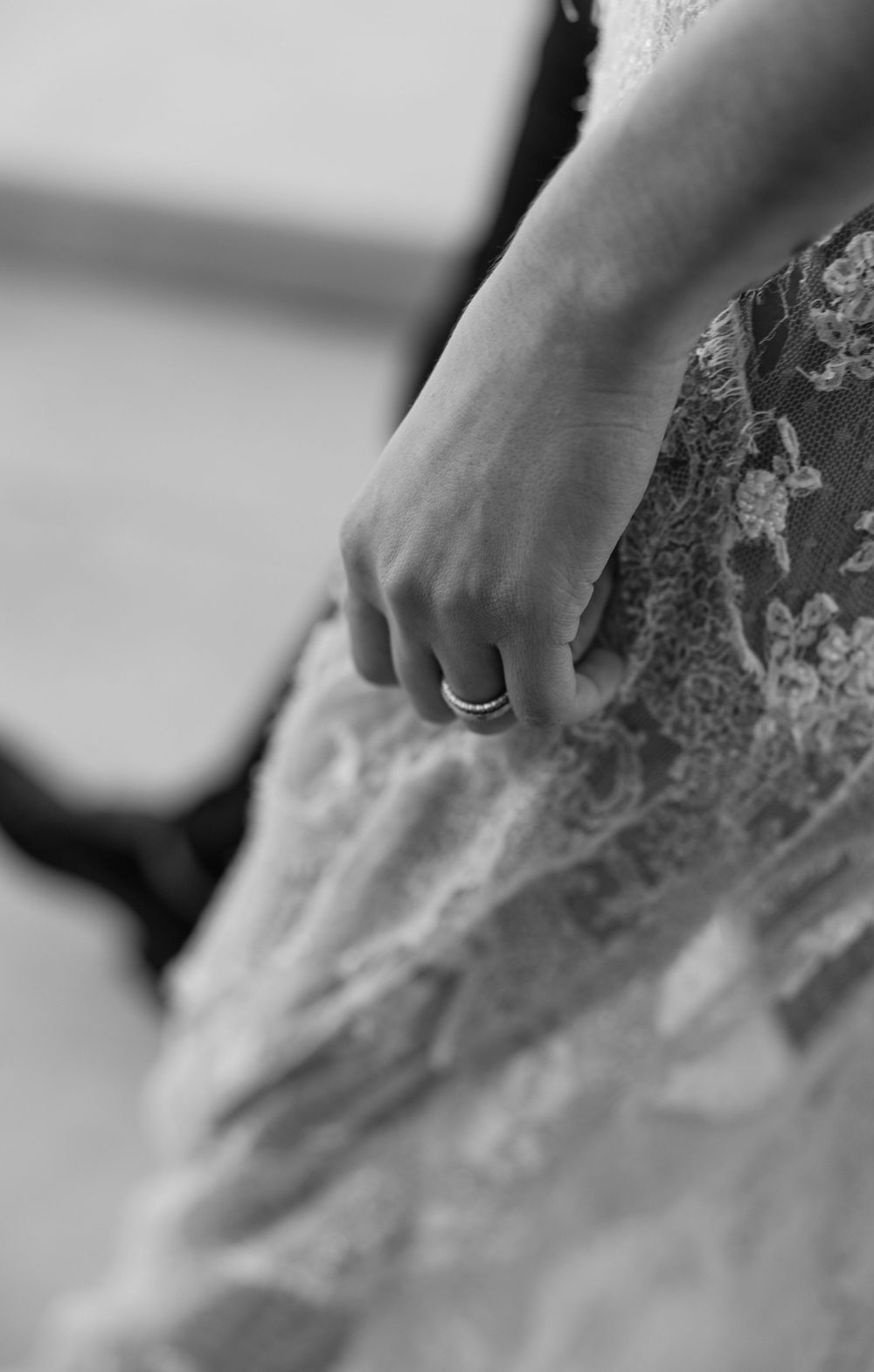 Close-up black and white photo of a delicate wedding ring on a hand against intricate lace fabric.