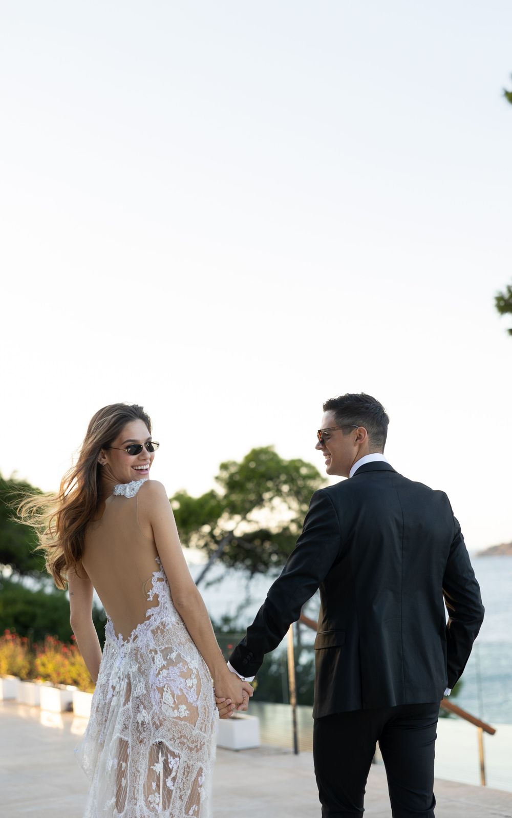 A romantic wedding couple holding hands while walking along a coastal path at dusk.