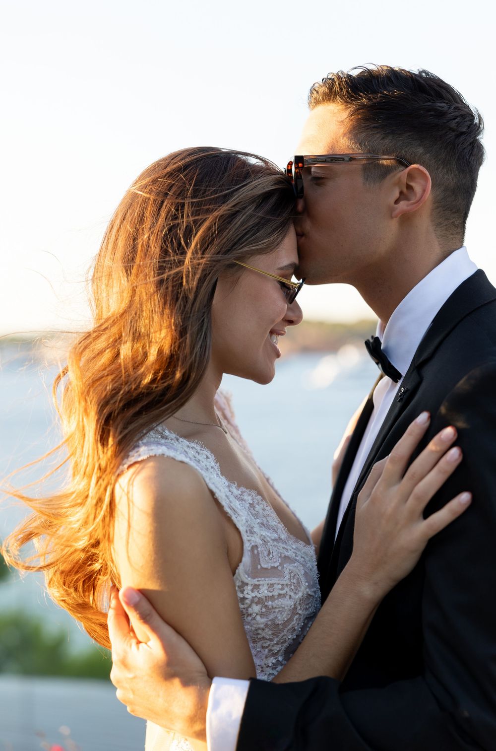 Romantic wedding portrait with golden sunlight illuminating couple against waterfront backdrop.