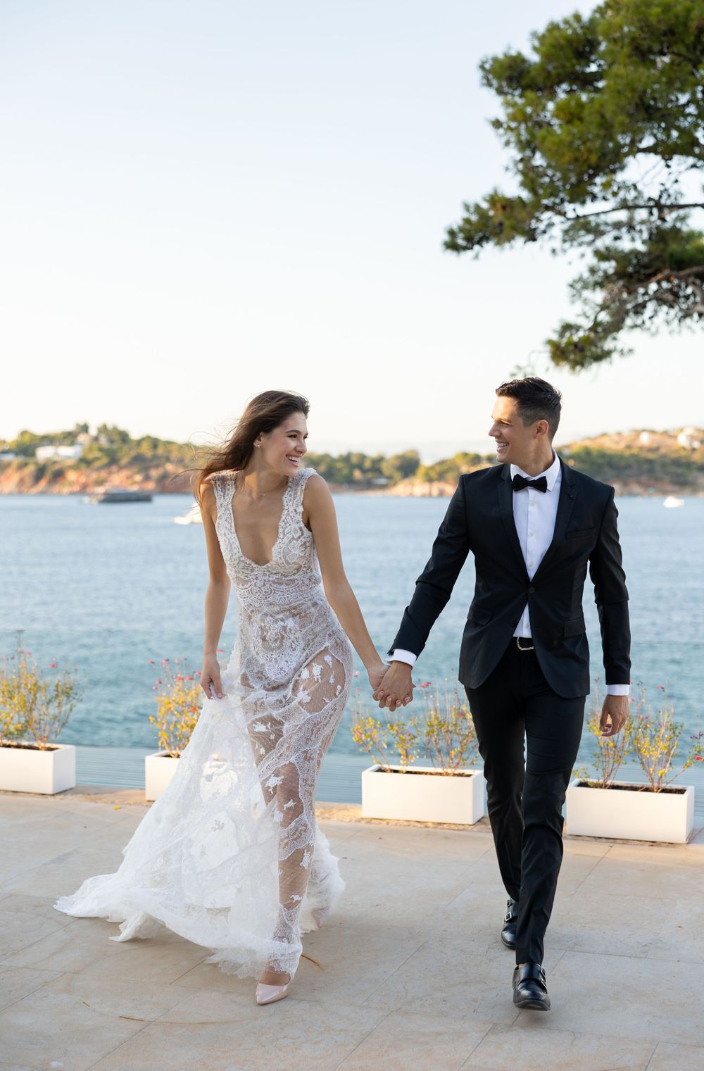 A newlywed couple walking hand in hand along a seaside terrace in formal wedding attire.