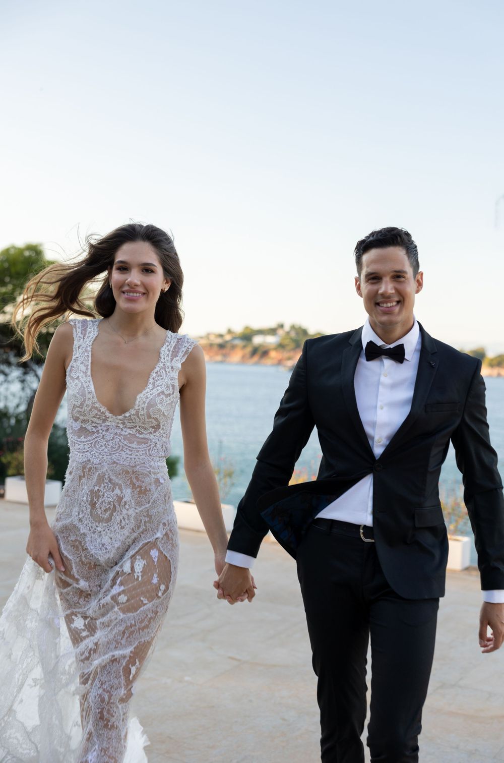 Joyful couple walking hand in hand on a seaside terrace in wedding attire.