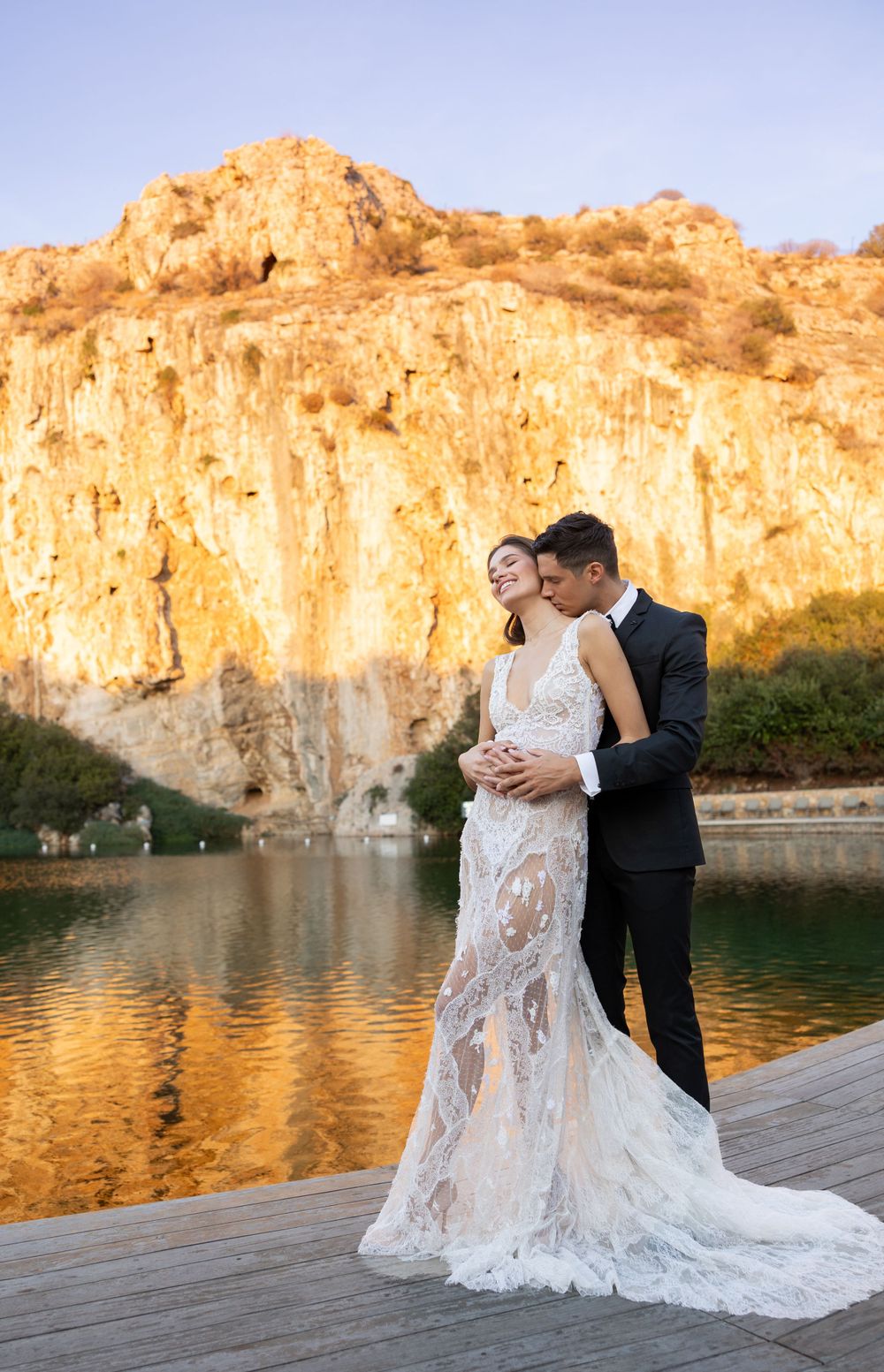 Romantic wedding photo with couple embracing on wooden deck by cliffside lake at golden hour.