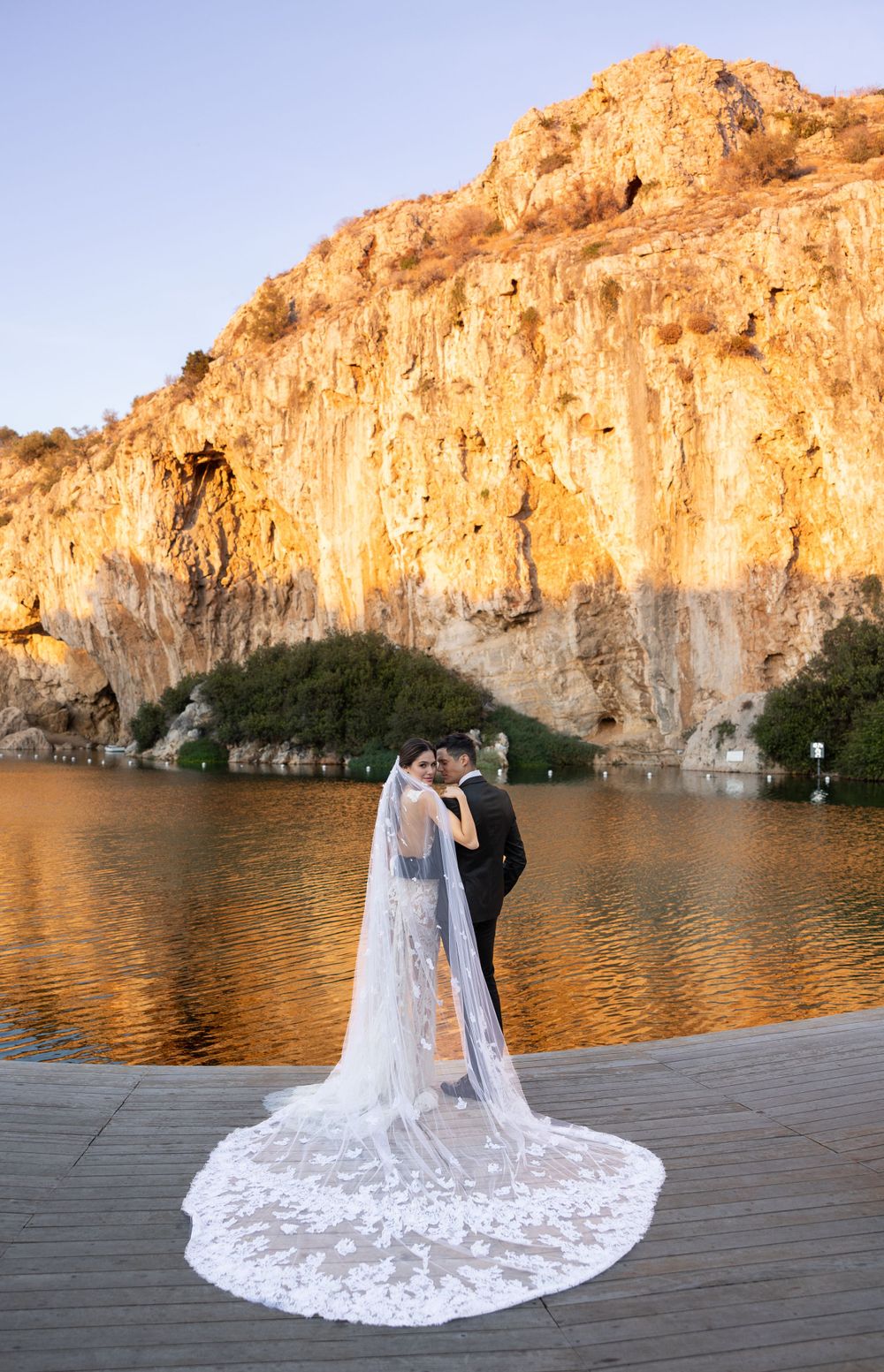 A wedding couple embracing on a dock with a dramatic golden cliff and reflective lake in the background.
