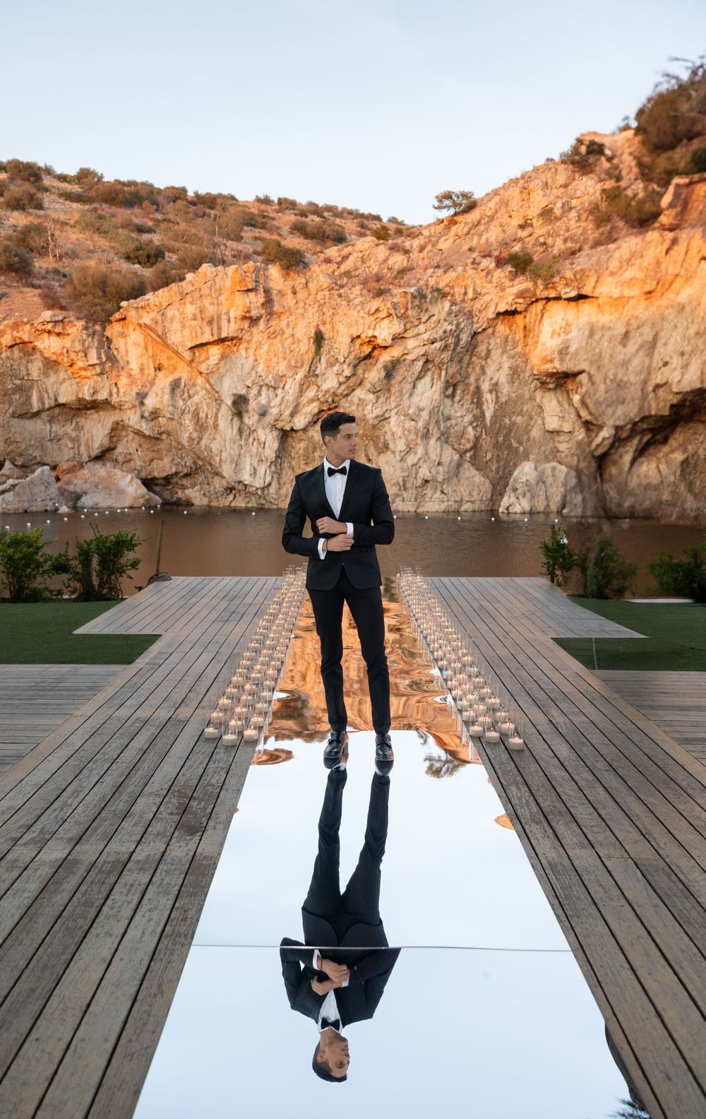 A black tuxedo-clad individual stands on a mirrored walkway with dramatic desert cliffs and glowing lights in the background.