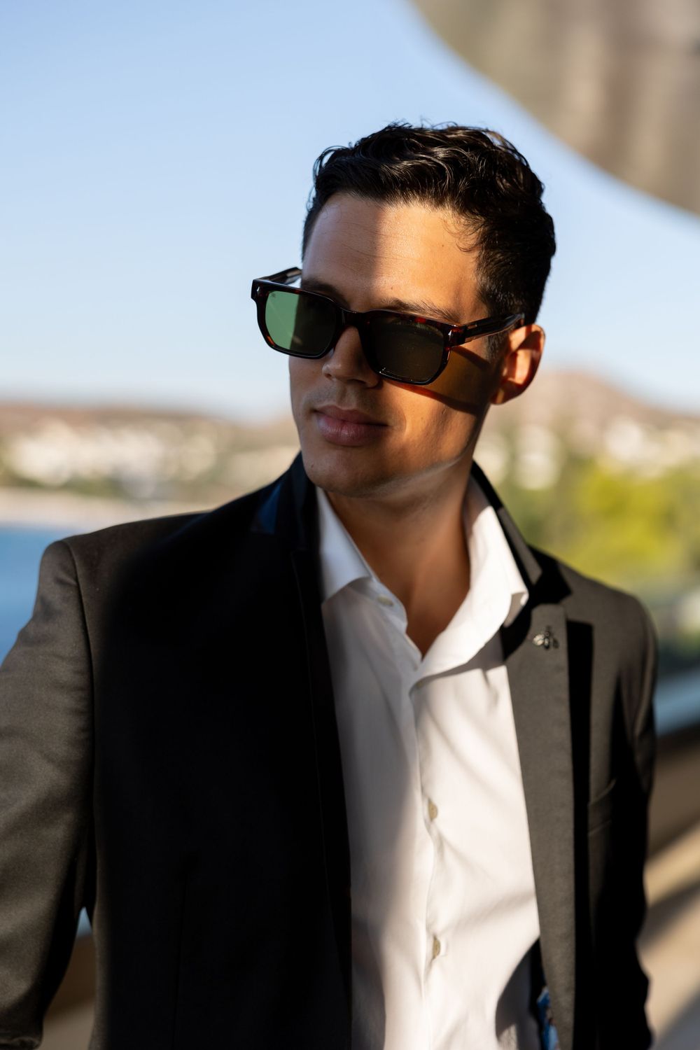 Stylish person in a black blazer and white shirt wearing sunglasses against a sunny waterfront backdrop.