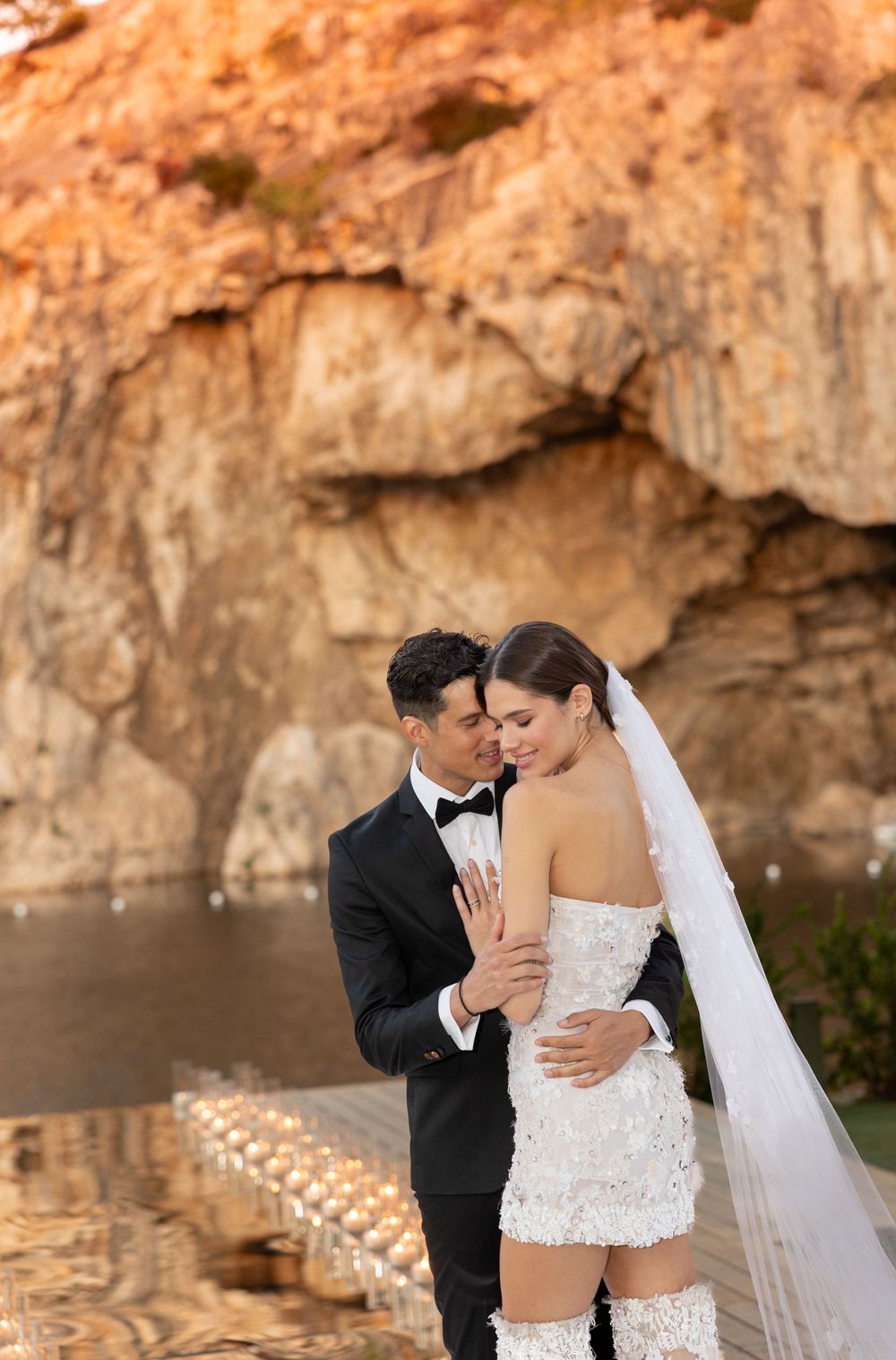 Couple in wedding attire embracing near rocky cliff face by seaside with flowing veil and short white dress.