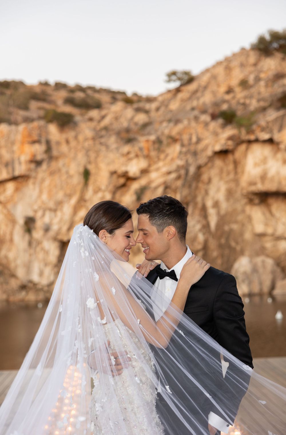 Intimate wedding moment captured on dock with dramatic cliff backdrop and flowing veil.