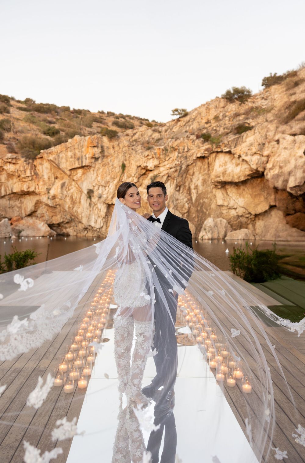 A wedding couple poses on a candlelit boardwalk against dramatic rocky cliffs at sunset.