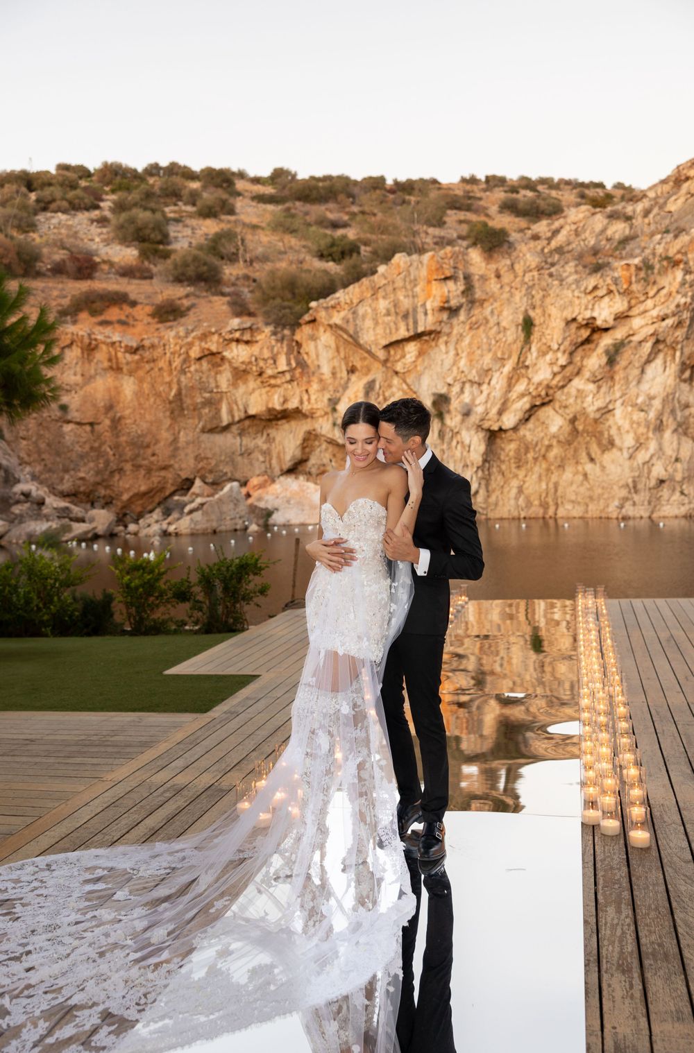 A couple embraces on a wooden deck with dramatic rocky cliffs forming a natural backdrop at sunset.