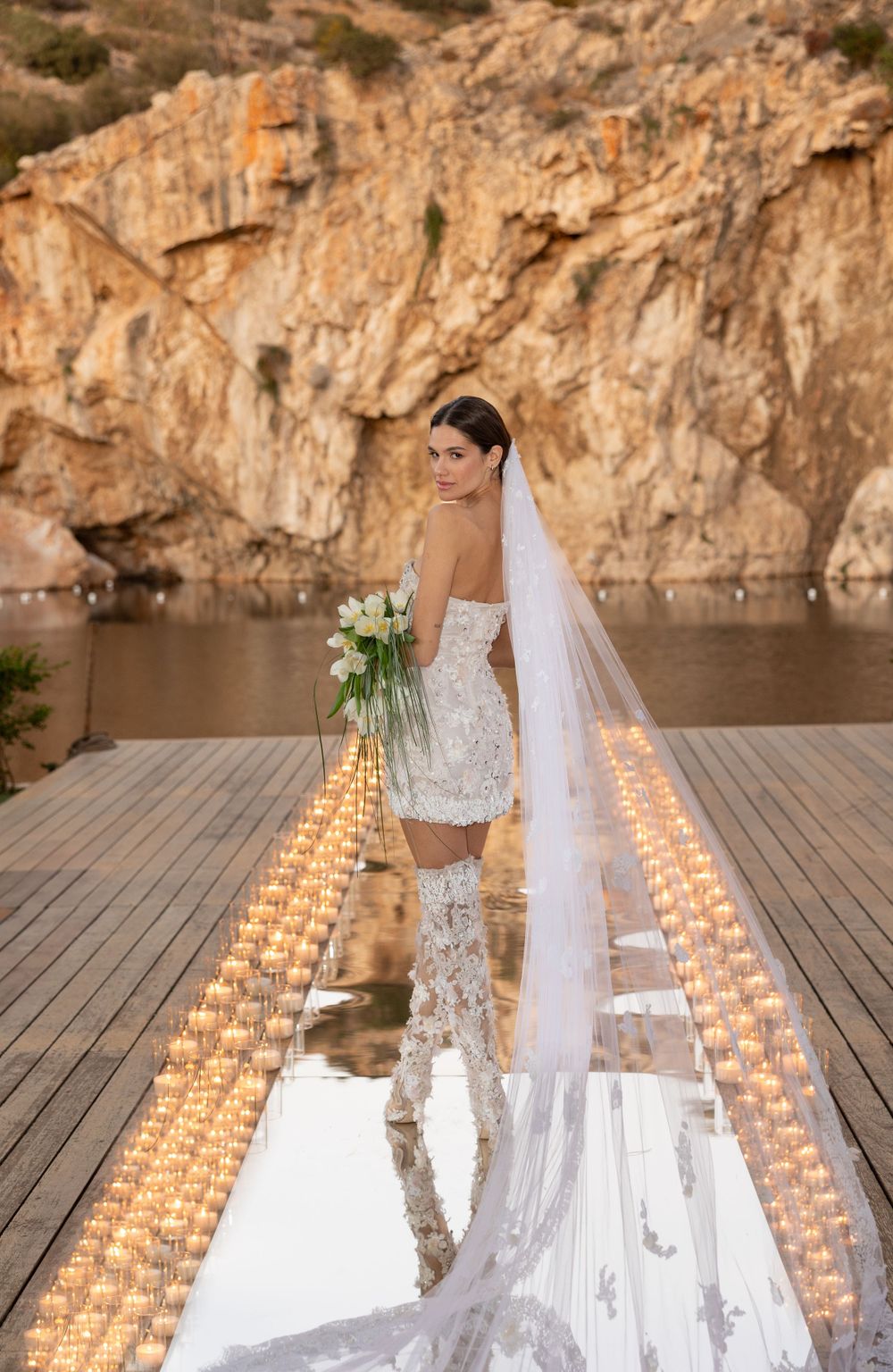 A bride in a lace dress poses on a wooden deck lined with candles against a rocky cliff backdrop.