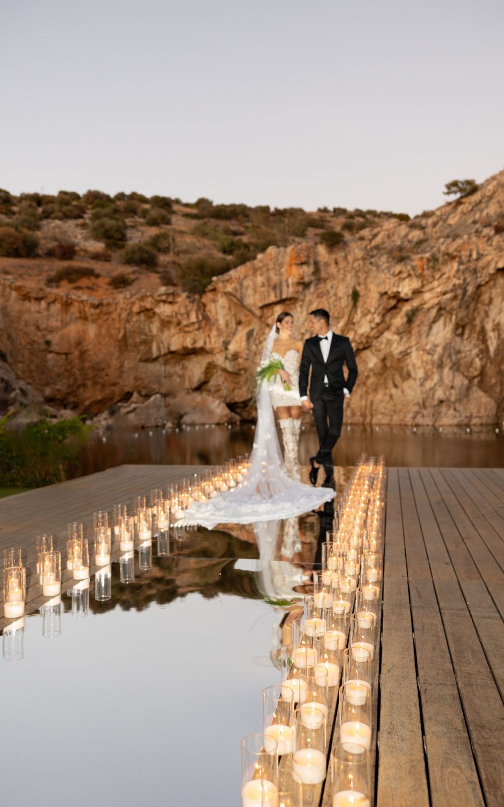 A romantic wedding scene on a wooden dock lined with glowing candles reflecting in still water against rocky cliffs at dusk.