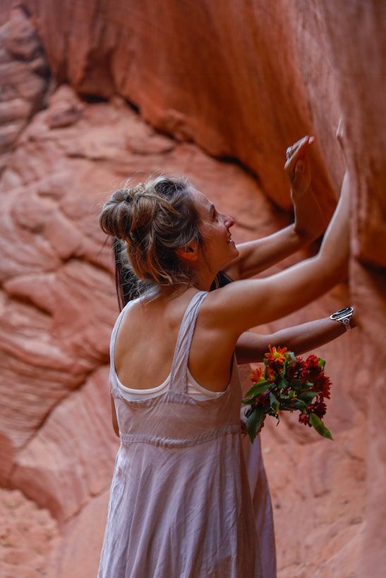 A couple of women in a flowing white dress holds red flowers while posing against red rock canyon walls.