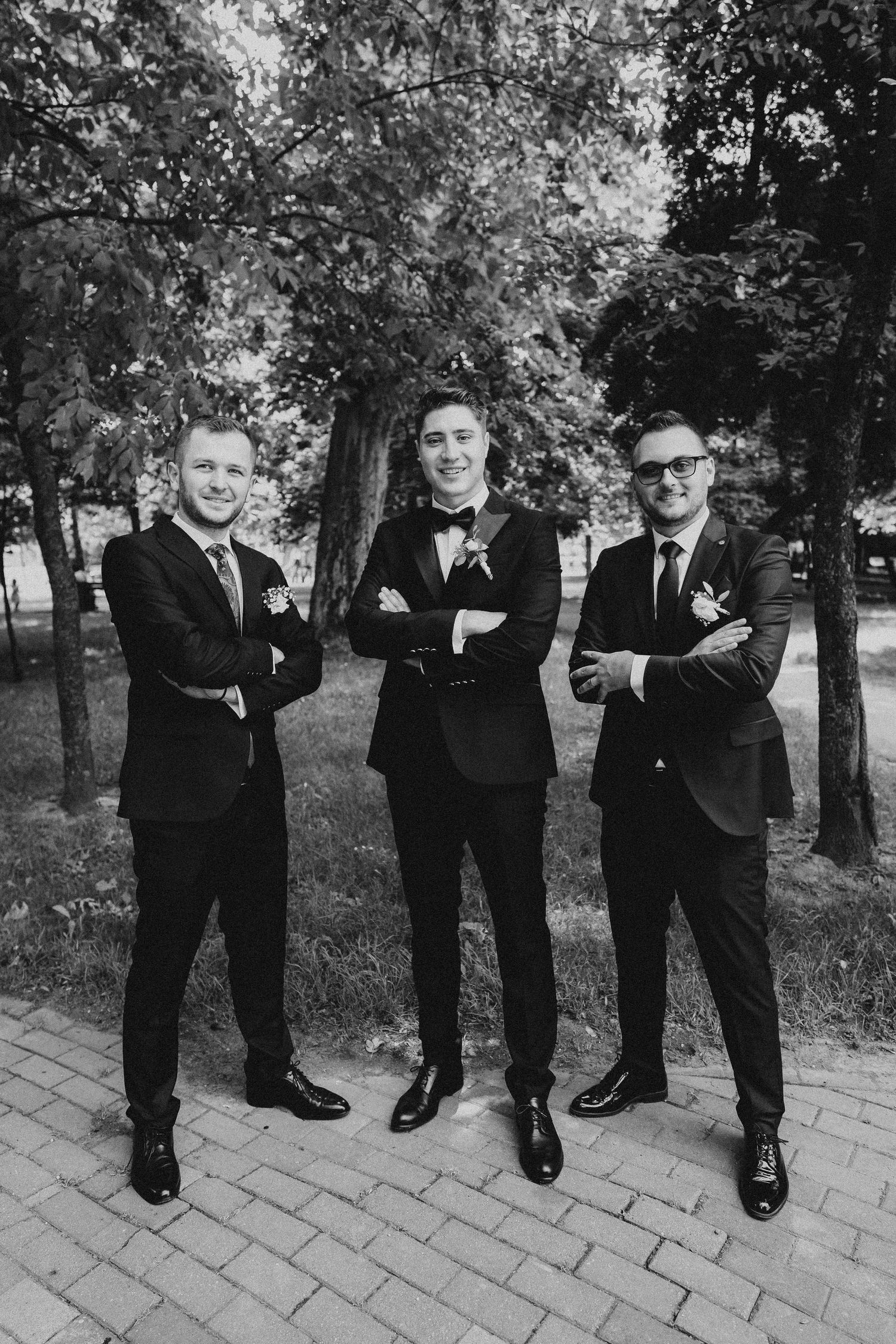 Groomsmen in black suits pose playfully on brick pavement during wedding photoshoot.