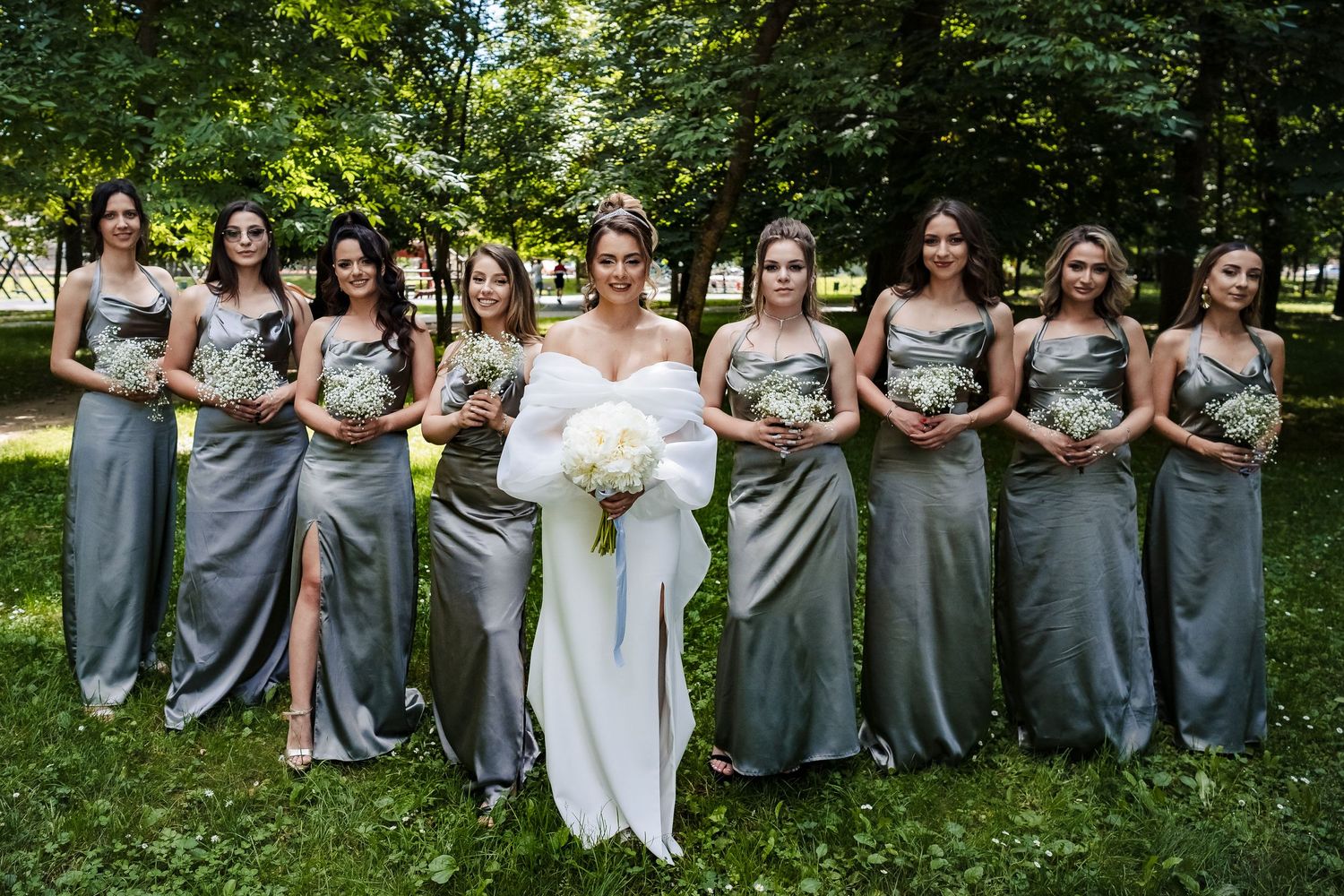 Wedding party in gray dresses poses outdoors with white bouquets against natural backdrop.