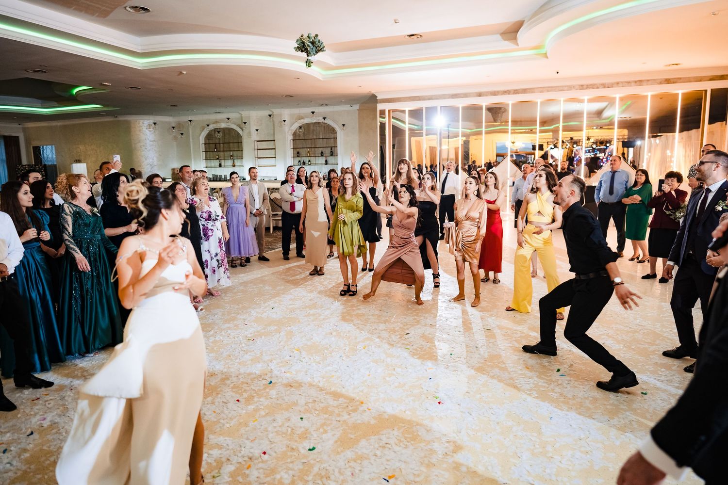 Guests watch excitedly during a wedding bouquet toss in an ornate ballroom with warm lighting.