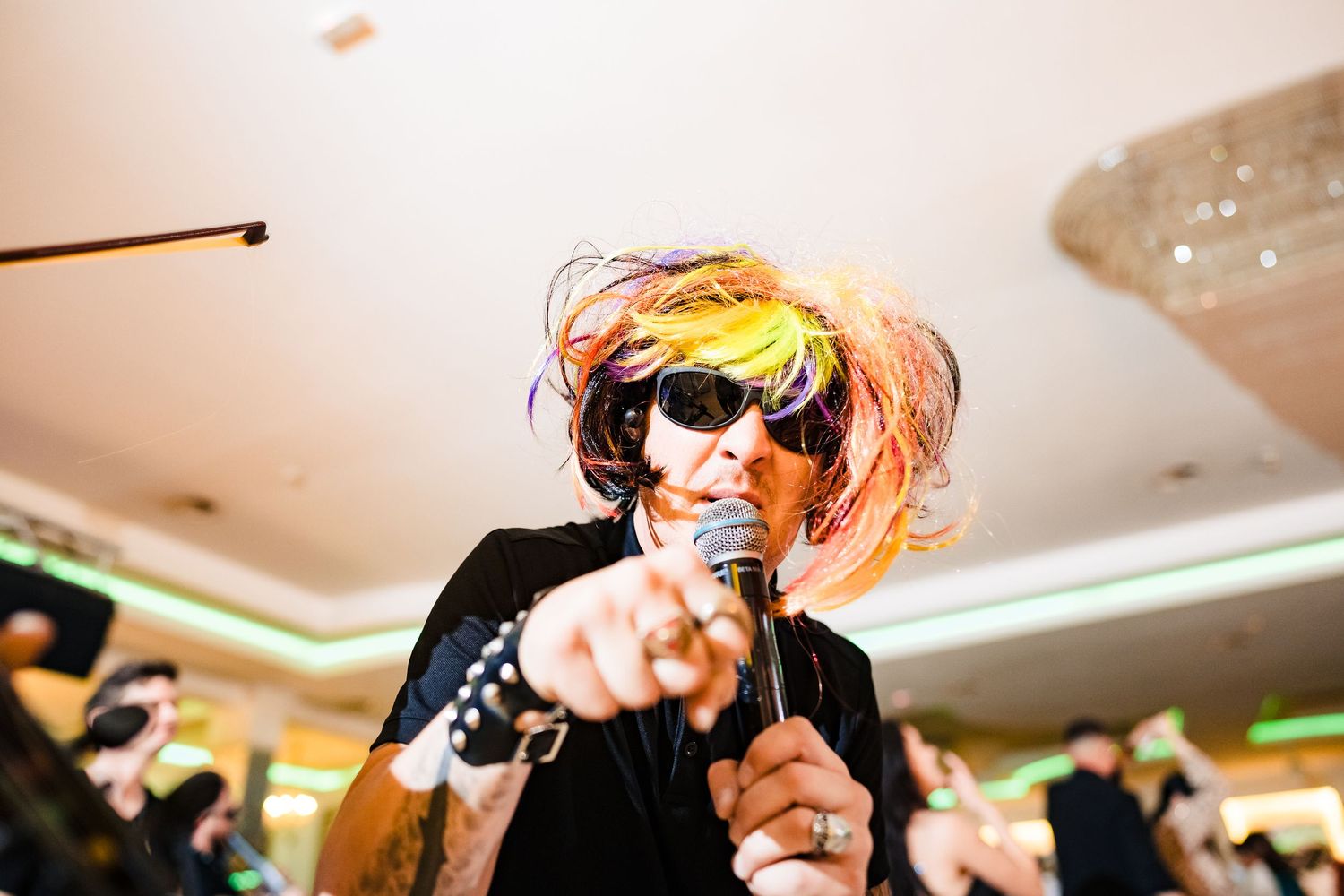 Performer in sunglasses and colorful wig sings into microphone during wedding entertainment.