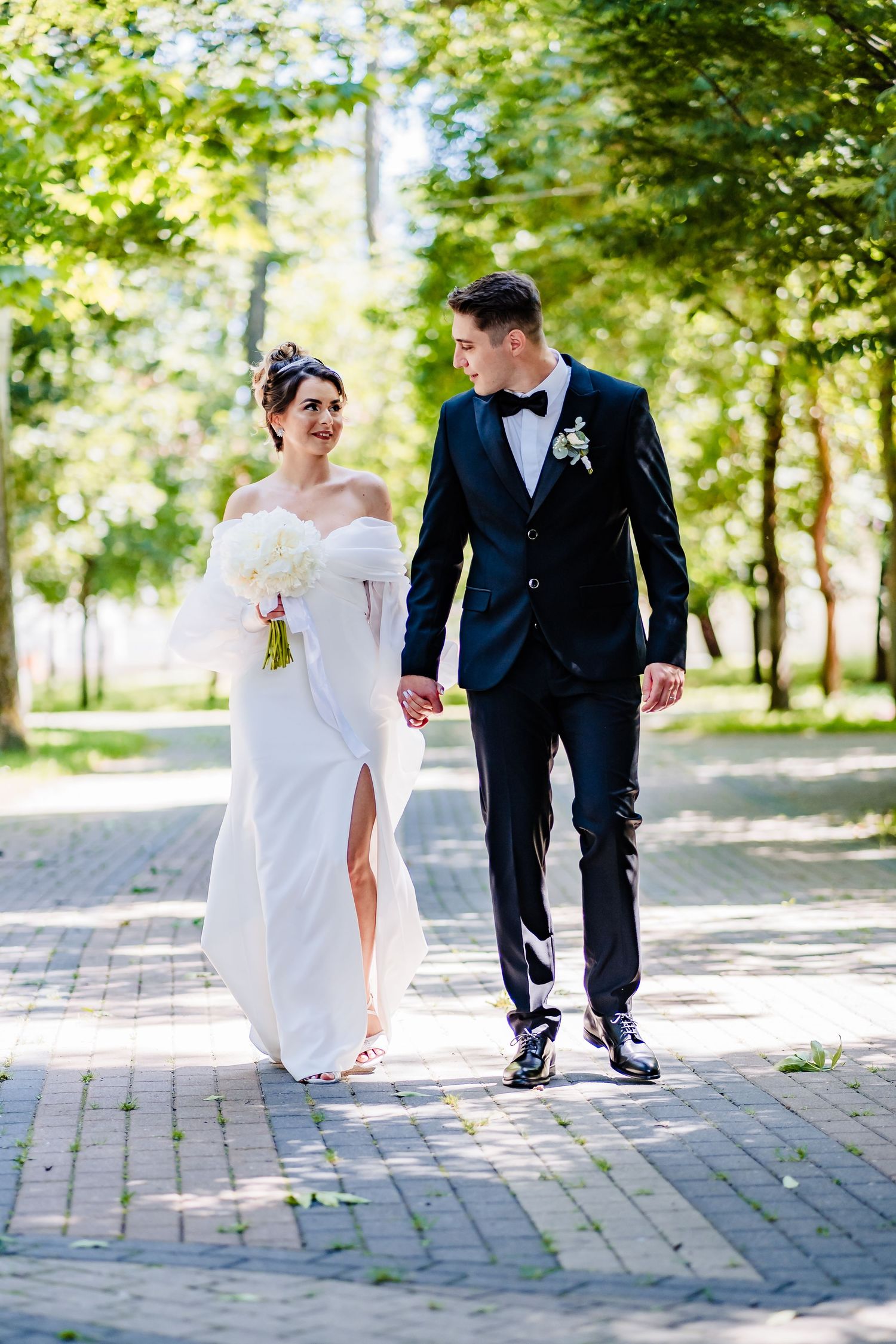 A bride in white dress walks with groom along a brick pathway surrounded by green trees.