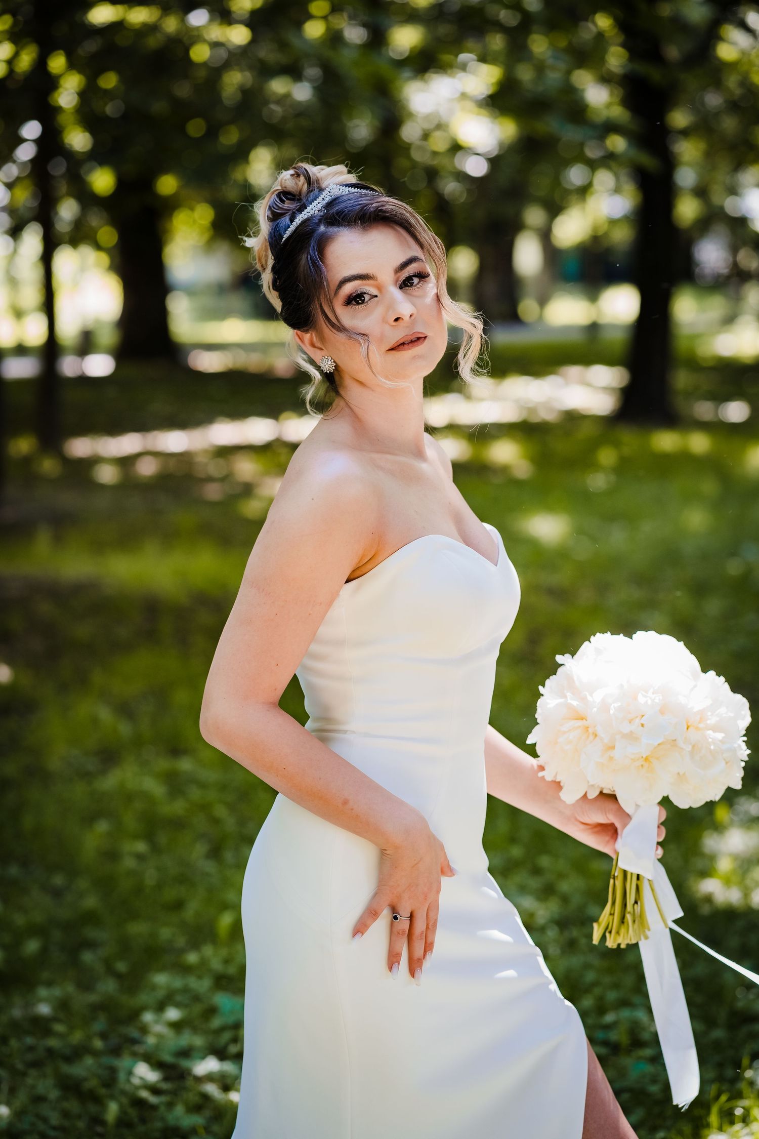 Bride in white strapless gown holding bouquet in outdoor garden setting.