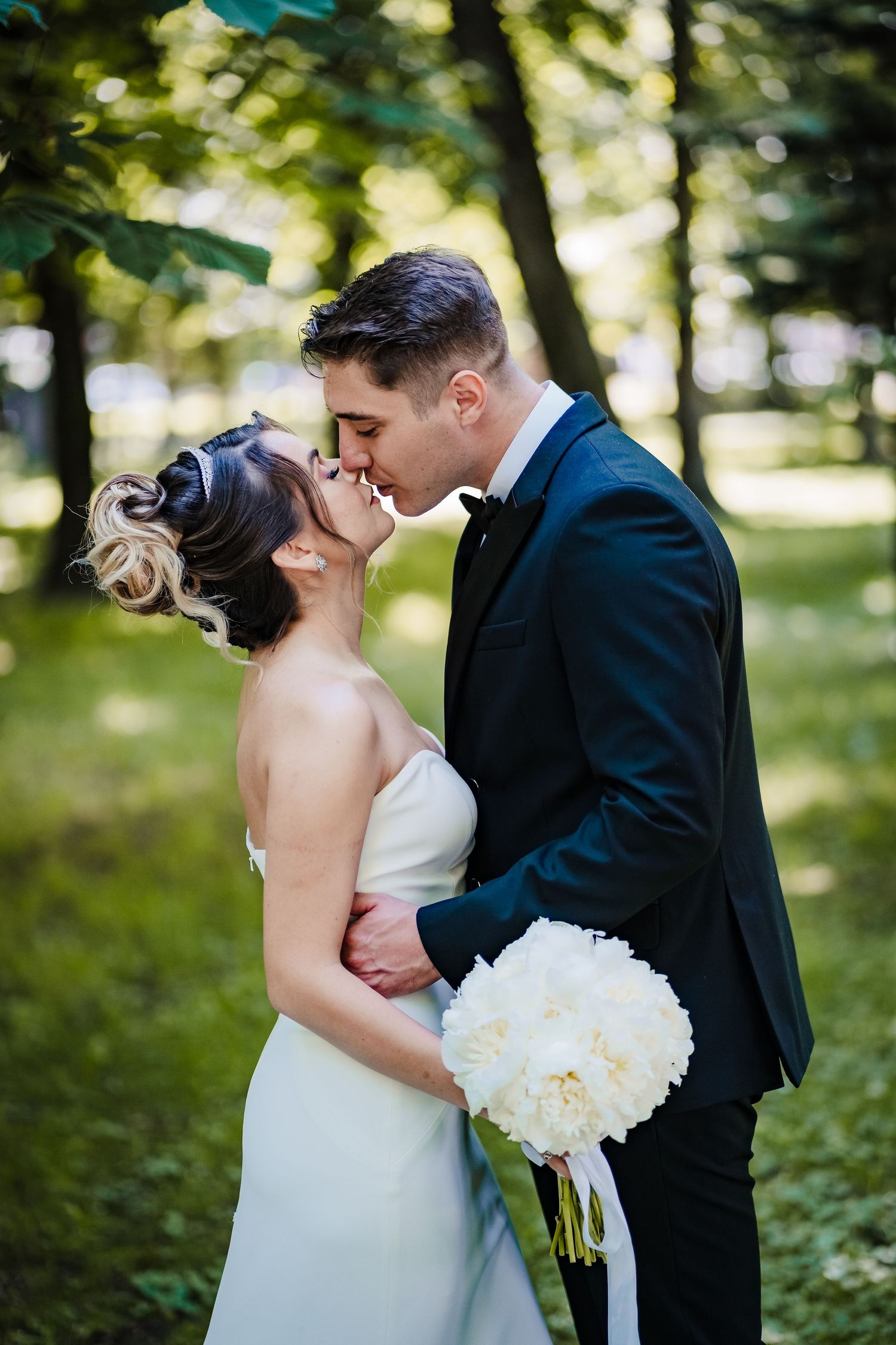 Elegant wedding couple embraces in a sun-dappled forest setting with white floral bouquet.