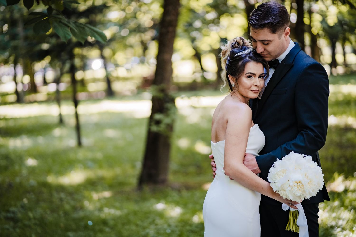 Wedding couple shares tender moments in sunlit forest with white floral bouquet.