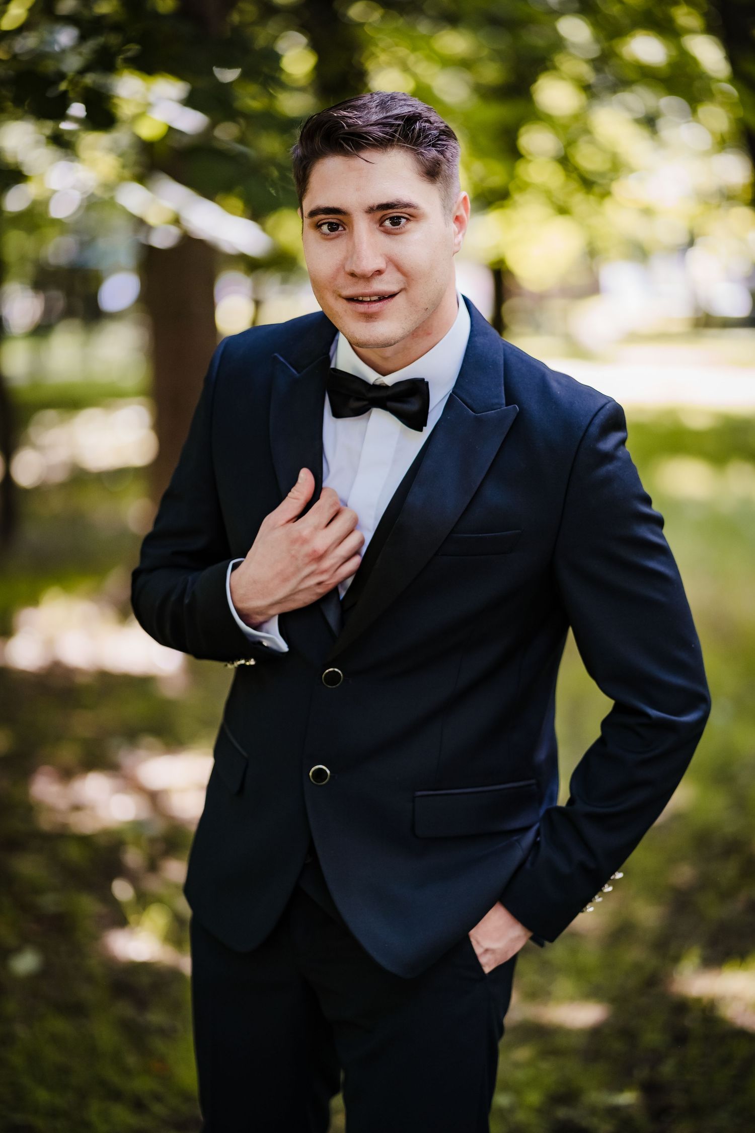 Groom in classic black tuxedo posing playfully in outdoor garden setting.